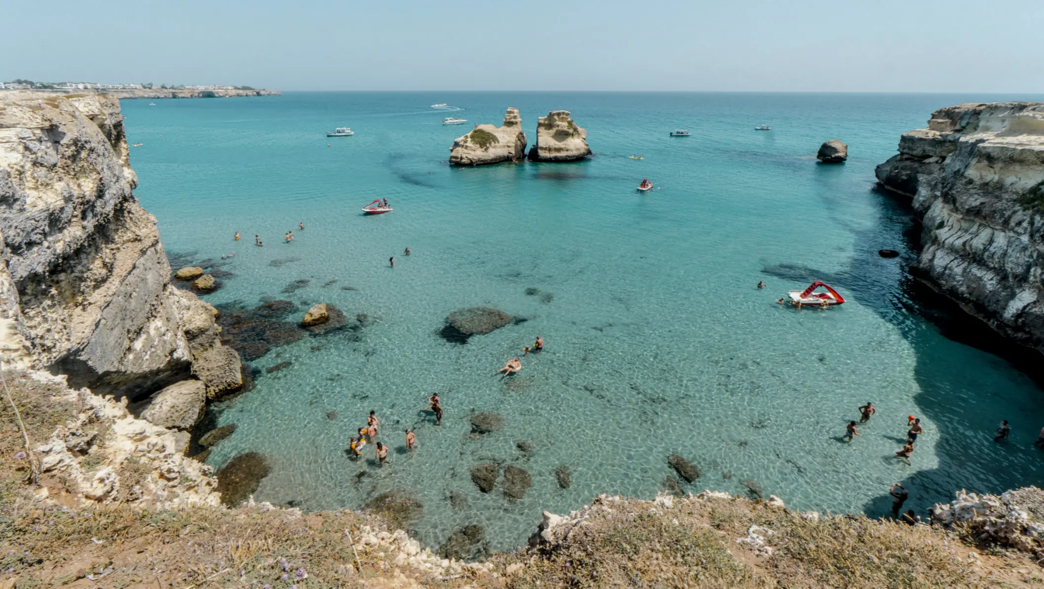 Türkisblaue Meeresbucht zwischen Felsklippen mit Schwimmern und kleinen Booten, zwei markante Felsformationen im Wasser.