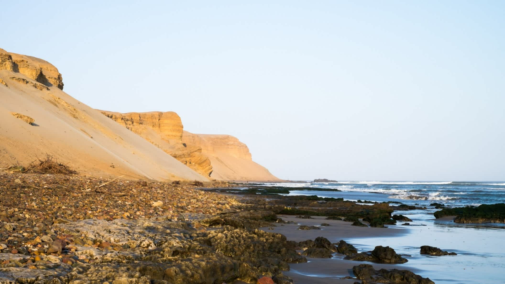 Blick auf den naturbelassenen wilden Strand mit Kieselsteinen am Strand und Sandbergen entlang der Küste sowie welligem Wasser.
