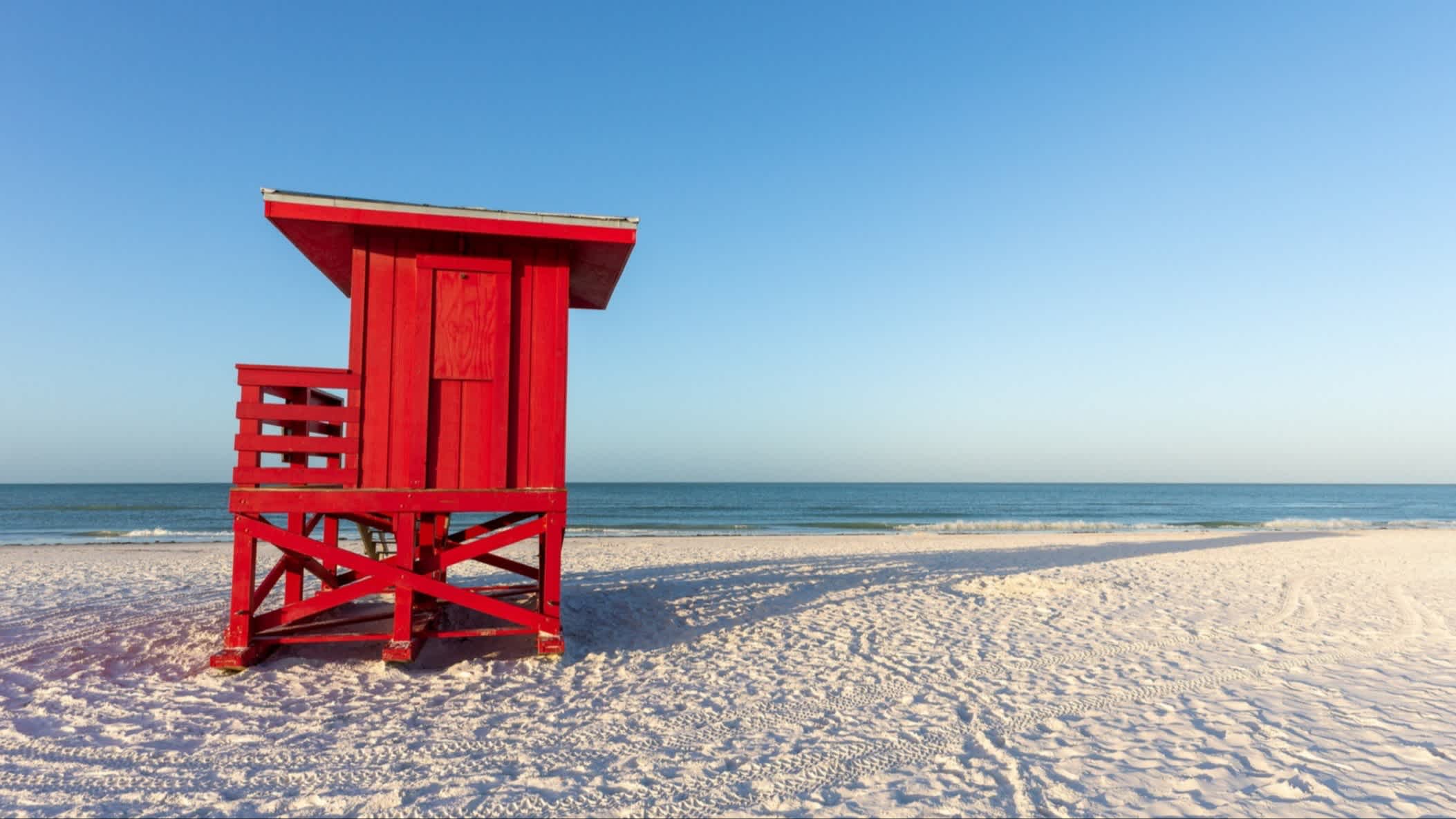 Der Strand Siesta Beach, Siesta Key, Florida, USA bei purem Sonnenschein mit Blick auf den weißen Sandstrand, das hellblaue Meer und einen feuerroten Wachturm aus Holz.