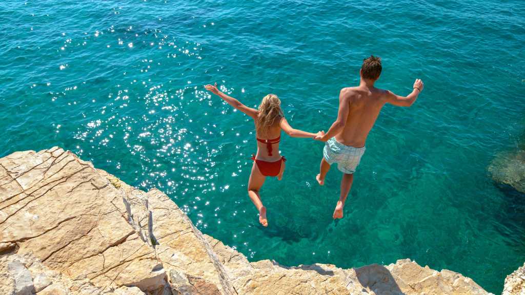 Zwei Personen springen von einer Klippe ins blaue Meer
