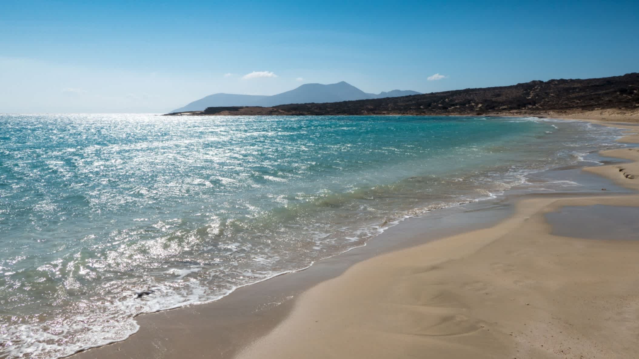 Der Pori Strand in Koufonisia, Kykladen, Griechenland mit Blick auf den goldenen Sand und das wellige blaue Meer sowie Bergen im Hintergrund.
