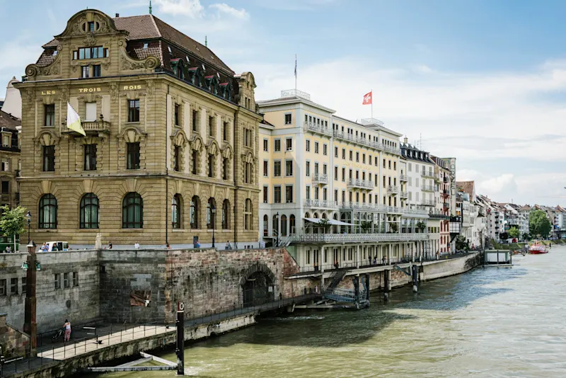 Historic buildings along a riverfront in Basel, Switzerland, featuring ornate architecture and a Swiss flag flying above.