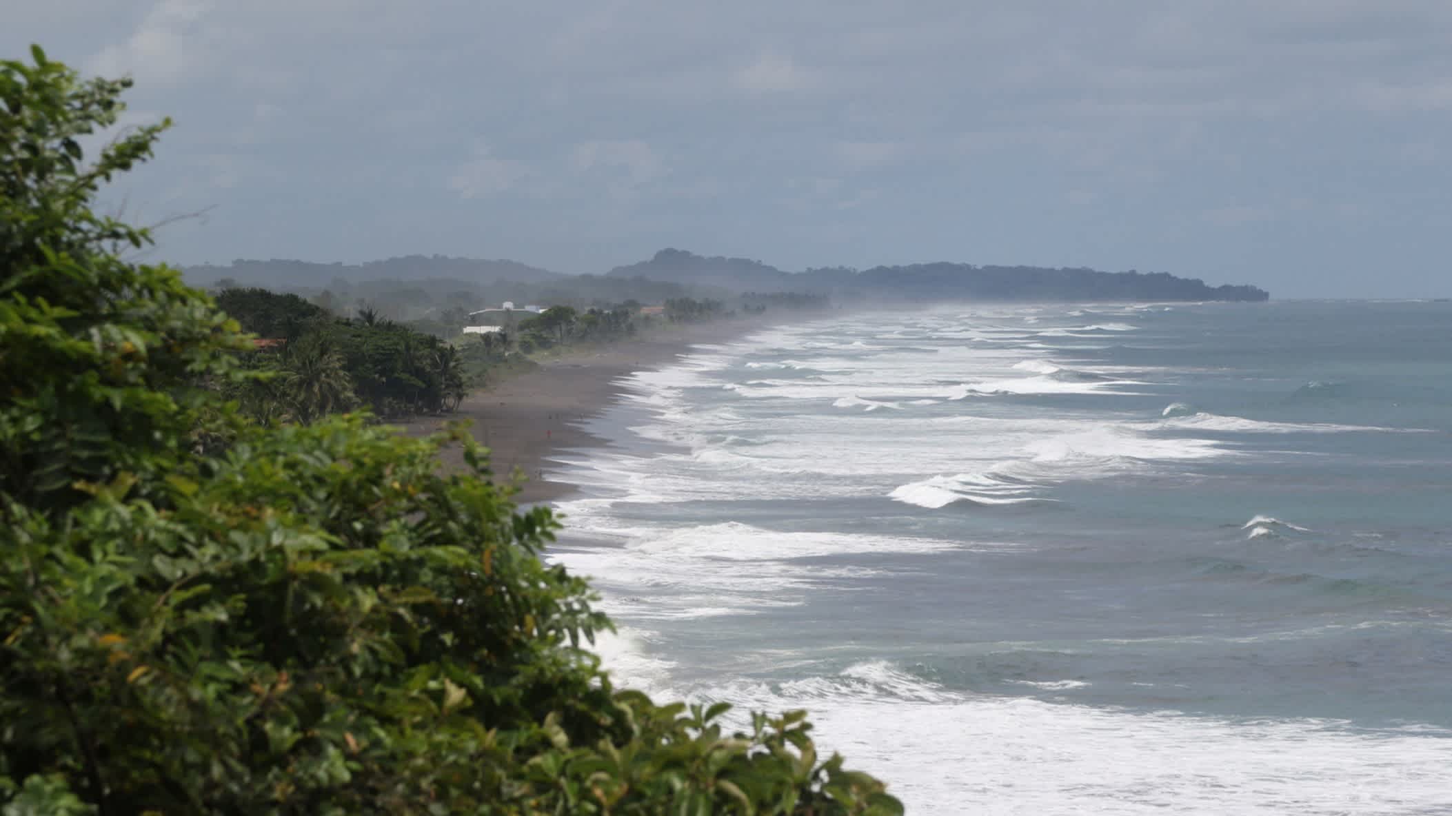 Die wunderschöne Pazifikküste in der Nähe von Jacó in Costa Rica mit welligem Meer, Dschungel und Bergen im Hintergrund bei bewölktem Himmel.


