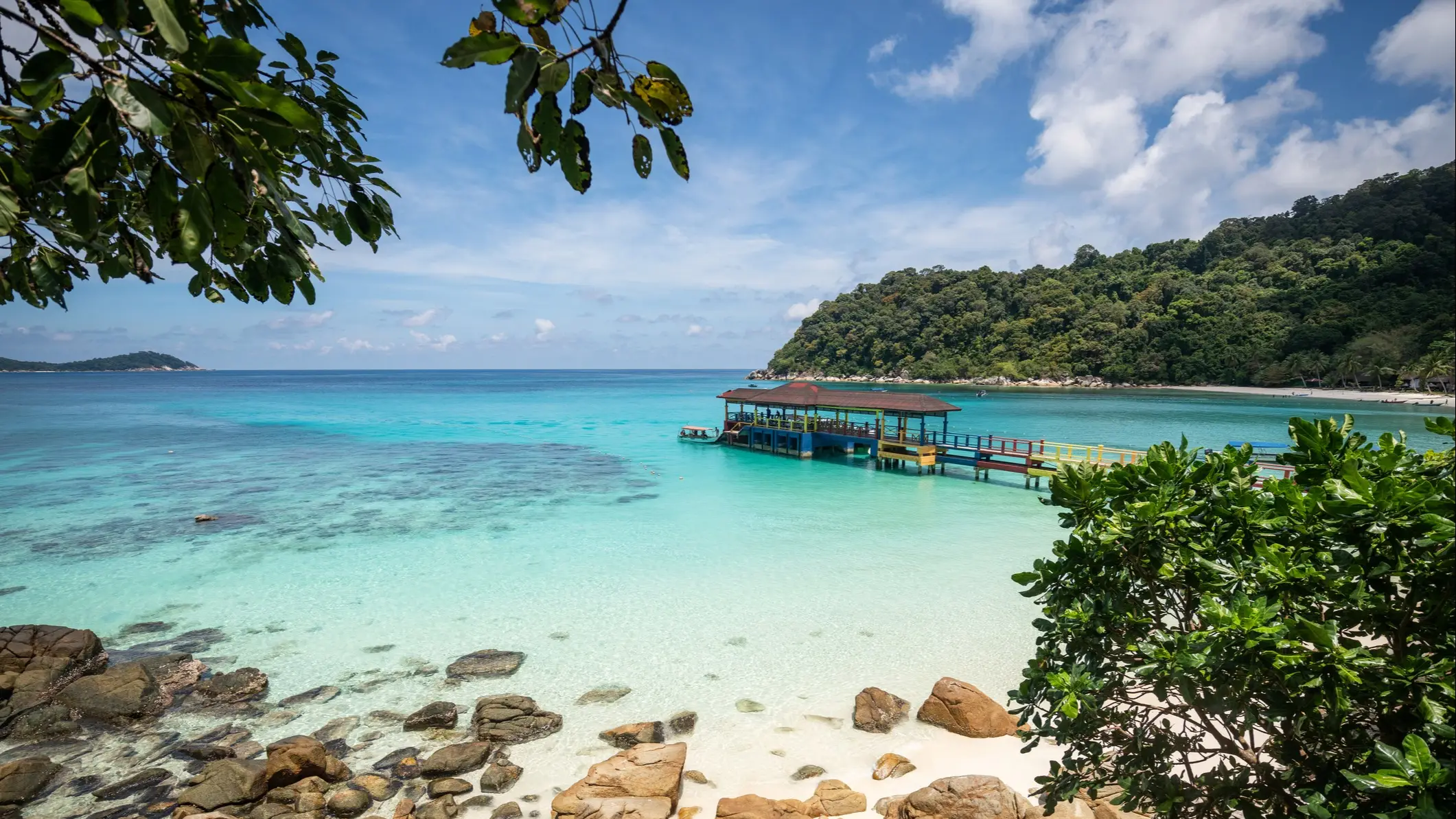 Türkisblaues Wasser mit Steg und Felsen, umgeben von üppiger Vegetation, Pulau Perhentian, Malaysia.
