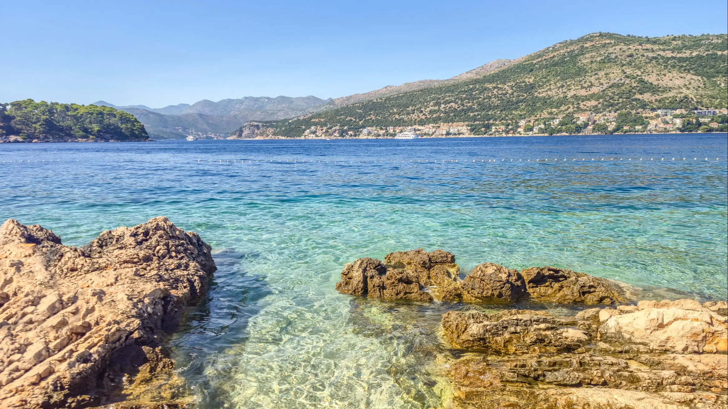 Kroatien Copacabana Der Strand von Copacabana-Strand, Dubrovnik, Kroatien mit Felsen und Häusern entlang der Bucht und Blick über das Wasser auf den Sonnenuntergang.