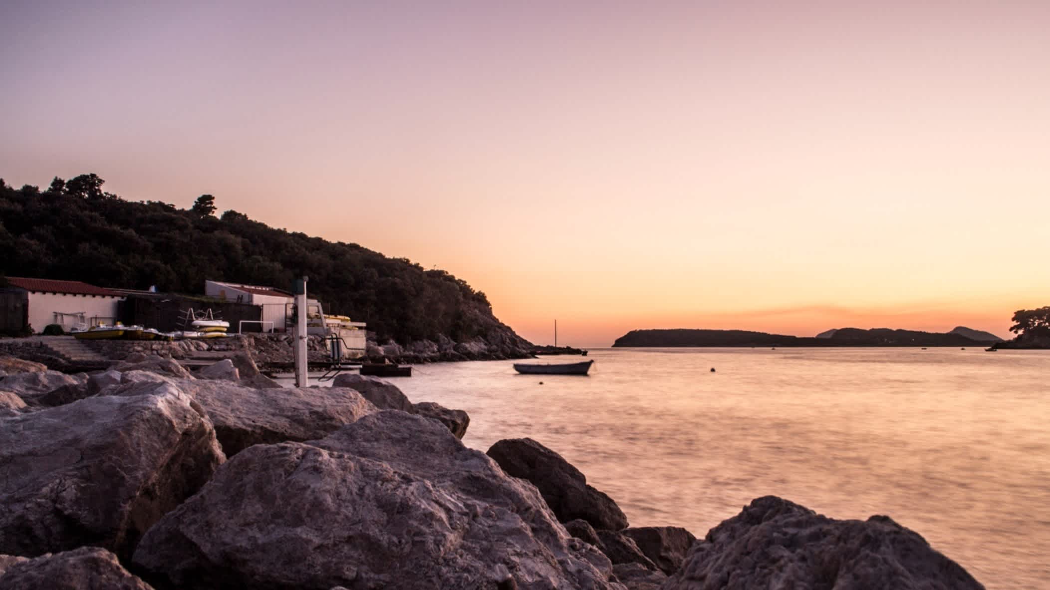 Der Strand von Copacabana-Strand, Dubrovnik, Kroatien mit Felsen und Häusern entlang der Bucht und Blick über das Wasser auf den Sonnenuntergang.