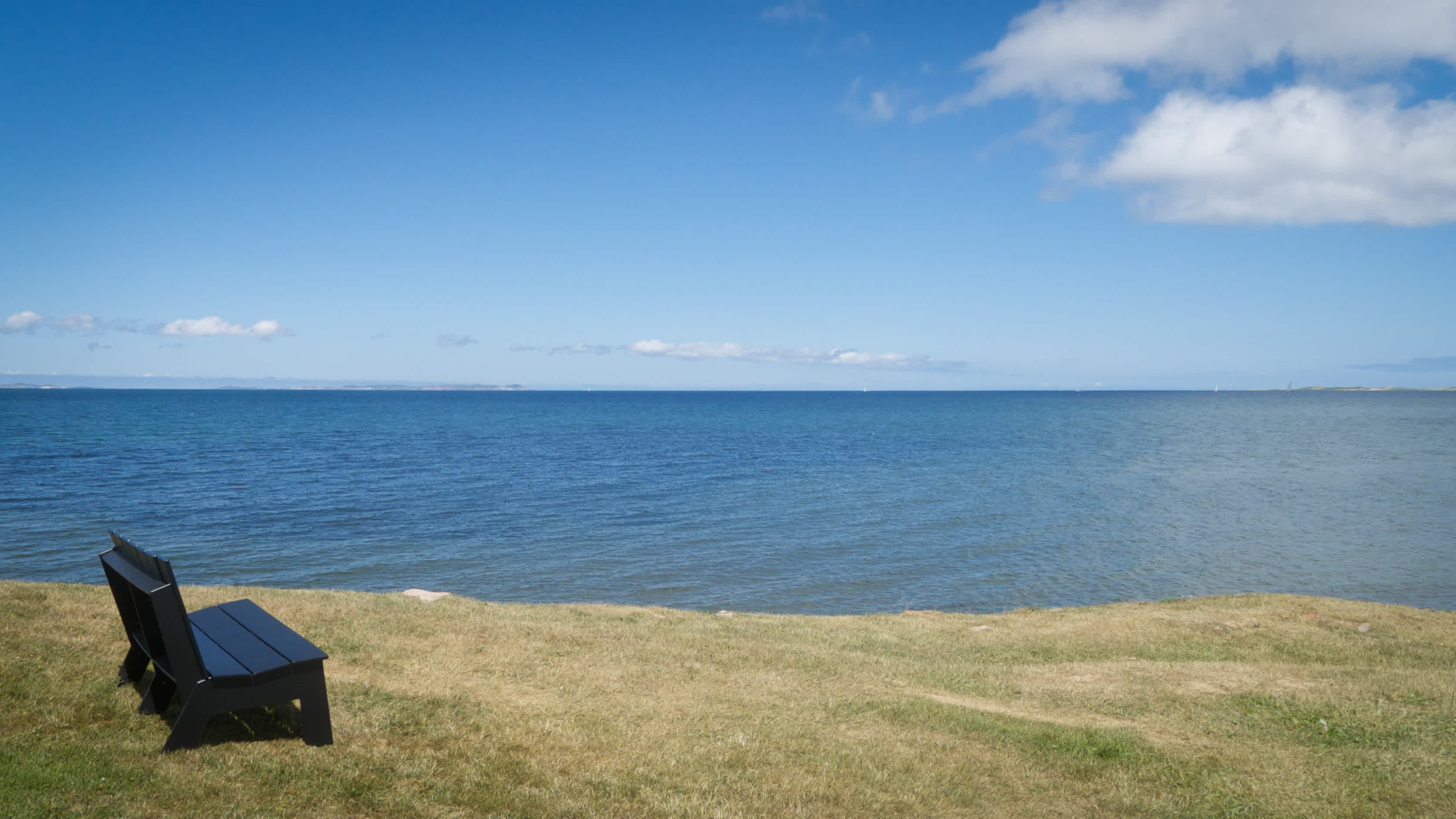 Eine Holzbank auf einer grünen Klippe am Strand Havre-Aubert Beach in Quebec, Kanada bei makellosem Sonnenschein und mit Blick auf das weite Meer.