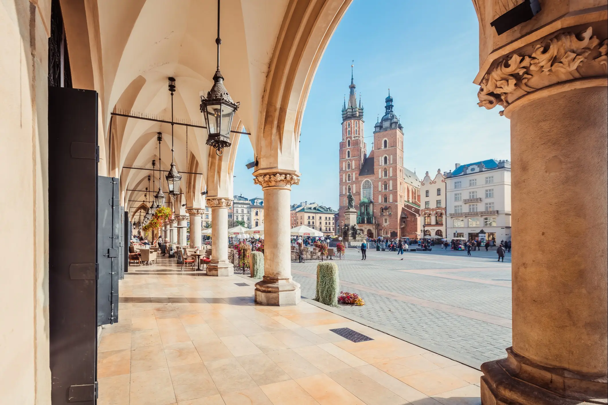 Blick durch Arkaden auf den Krakauer Hauptmarkt mit der Marienkirche und ihren charakteristischen Türmen bei blauem Himmel.