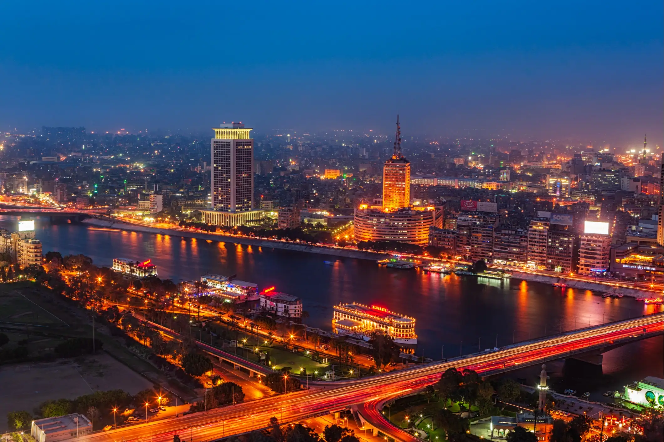 Nächtliche Skyline von Kairo mit beleuchteten Wolkenkratzern am Nil, Booten und Straßen mit Lichtspuren in der Dämmerung.