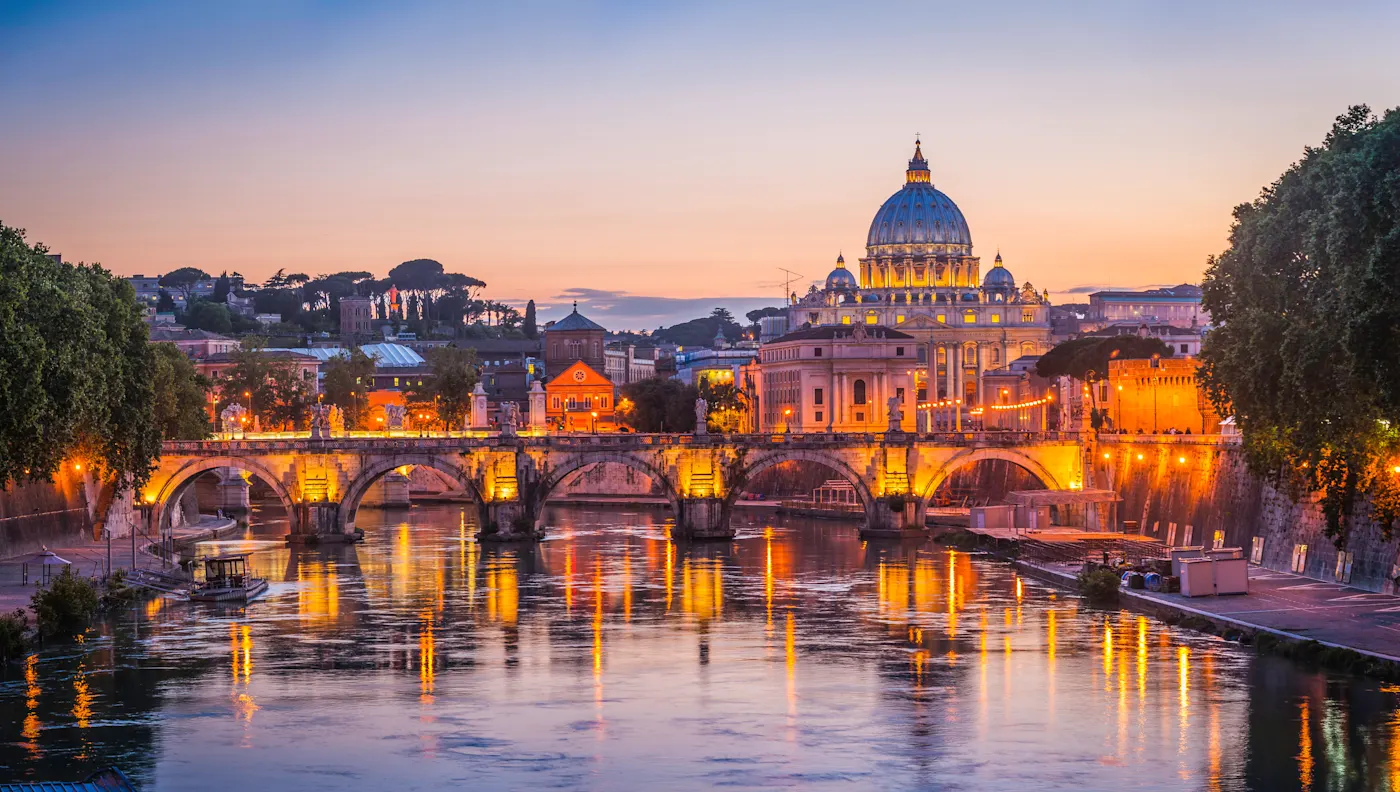 Italy_Rome_St Peters Basilica_sunset_iStock Ein malerischer Blick auf den Petersdom und den Vatikan bei Sonnenuntergang, mit warmen Lichtern, die die Brücke beleuchten und sich auf dem Tiber in Rom, Italien, spiegeln. Bäume umrahmen die Flussufer unter einem farbenfrohen Abendhimmel.