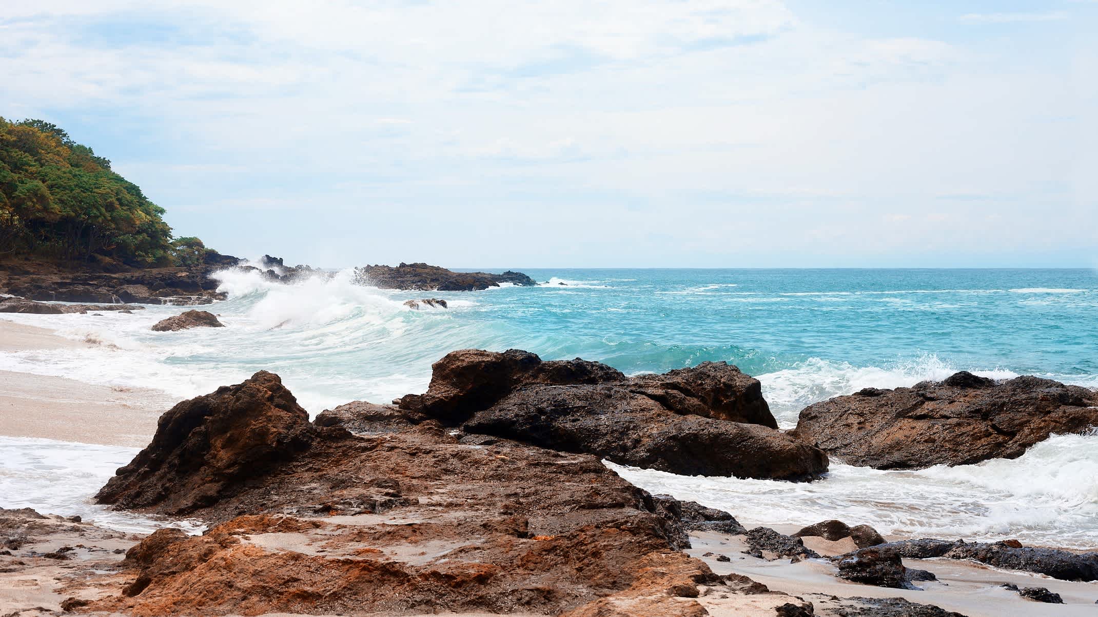 Der Strand von Montezuma in Costa Rica mit imposanten Wellen im blauen Meer und Felsen am weißen Sandstrand. 
