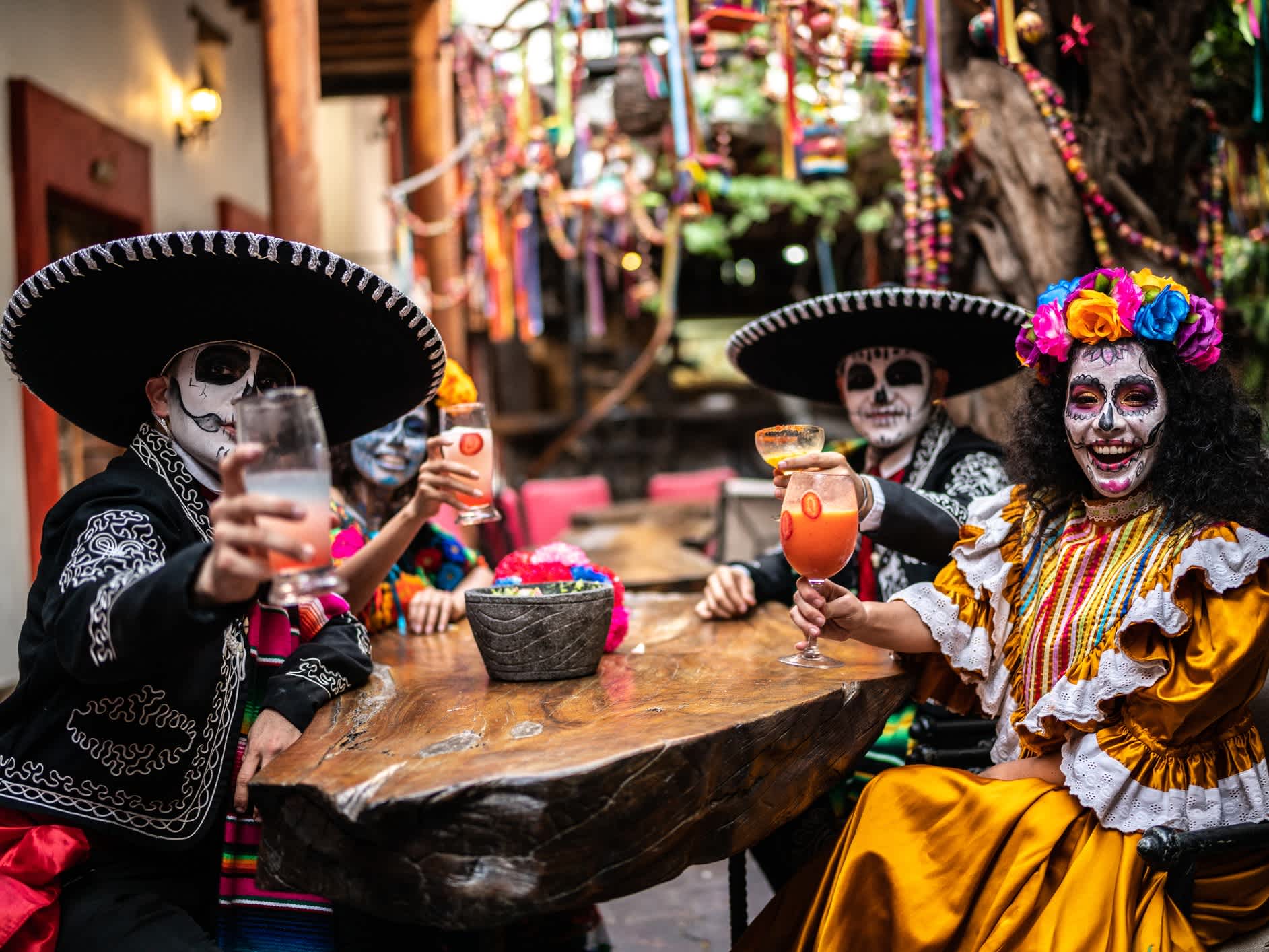 4 tanzende Menschen tragen bunte Trachten und ein Totenkopf-Make-Up zu Ehren des Día de los Muertos