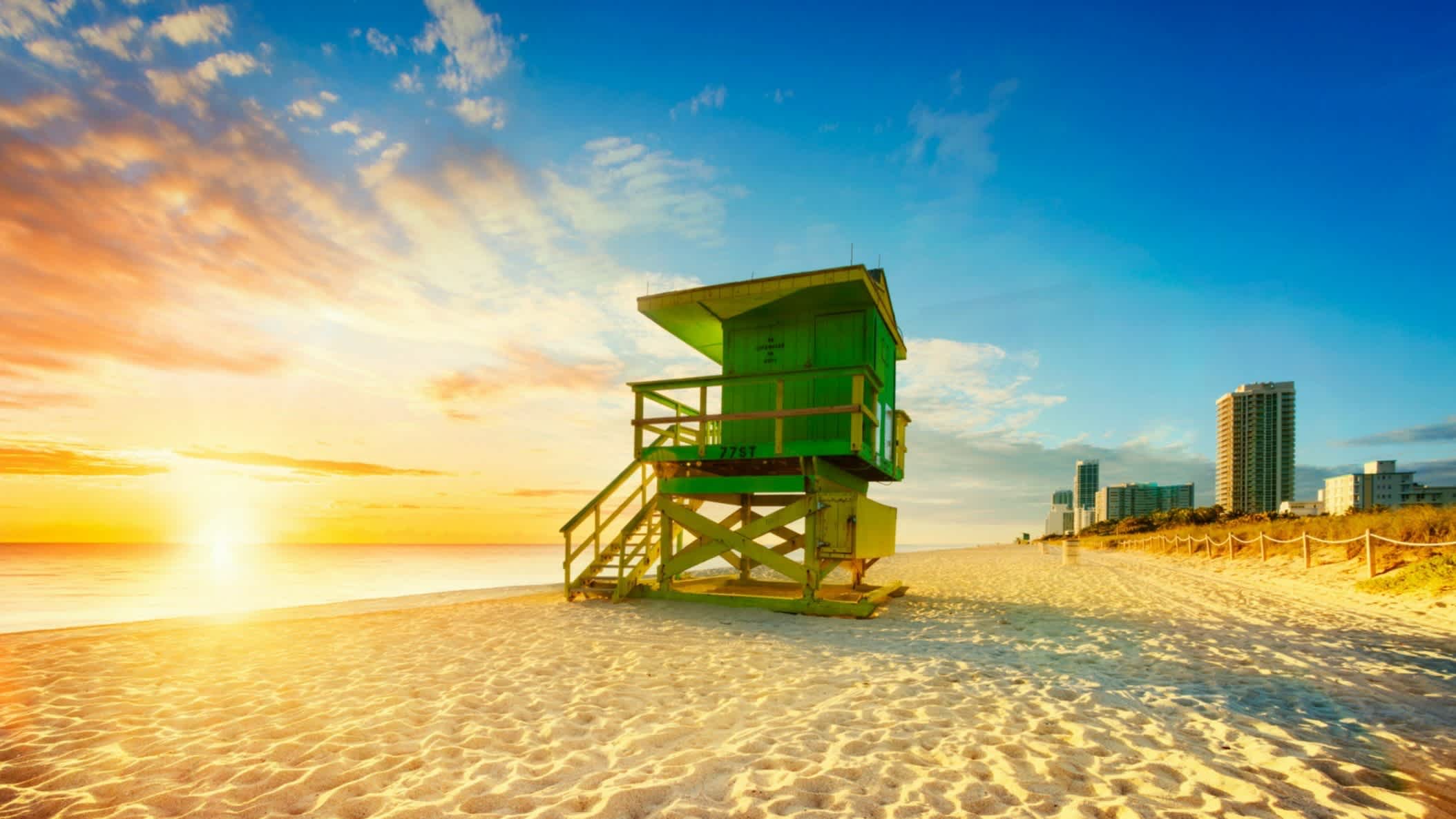 Der Strand South Beach, Miami Beach, Florida, USA bei farbenprächtigem Sonnenuntergang mit Blick auf einen grünen Wachturm sowie Hochhäusern im Hintergrund.