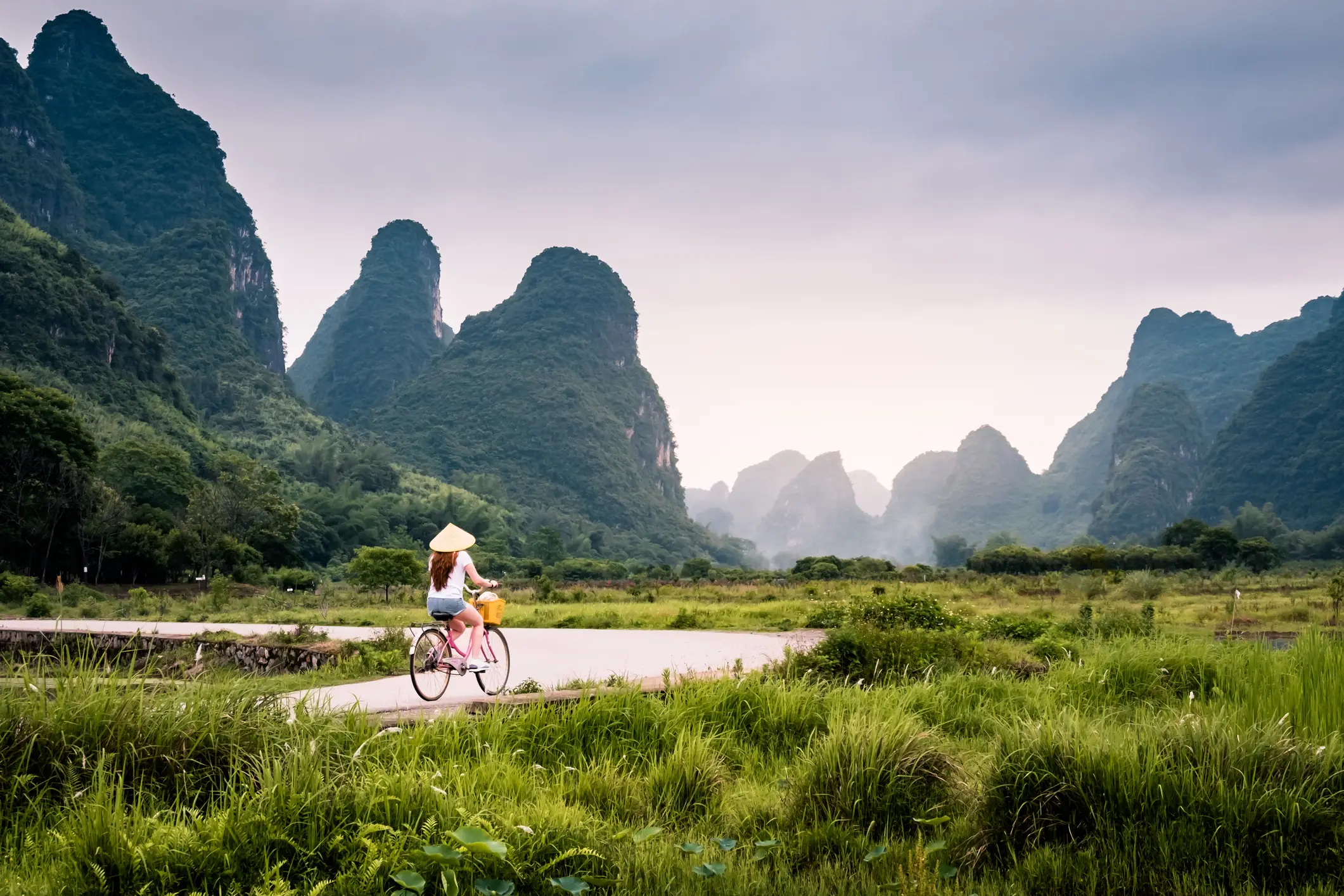 Person mit Strohhut fährt Fahrrad auf einem Pfad durch grüne Landschaft mit dramatischen Karstbergen in China.