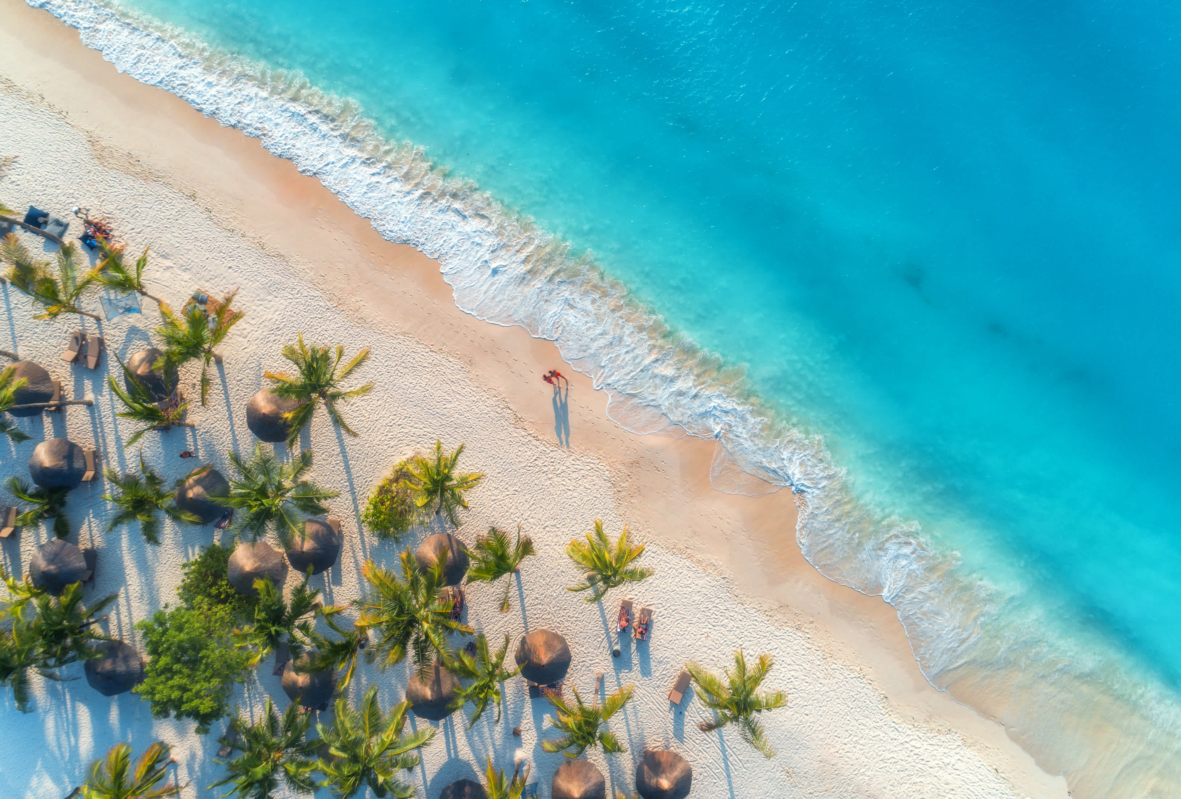 Luftaufnahme von Sonnenschirmen, Palmen am Sandstrand, Menschen, blaues Meer mit Wellen bei Sonnenuntergang
