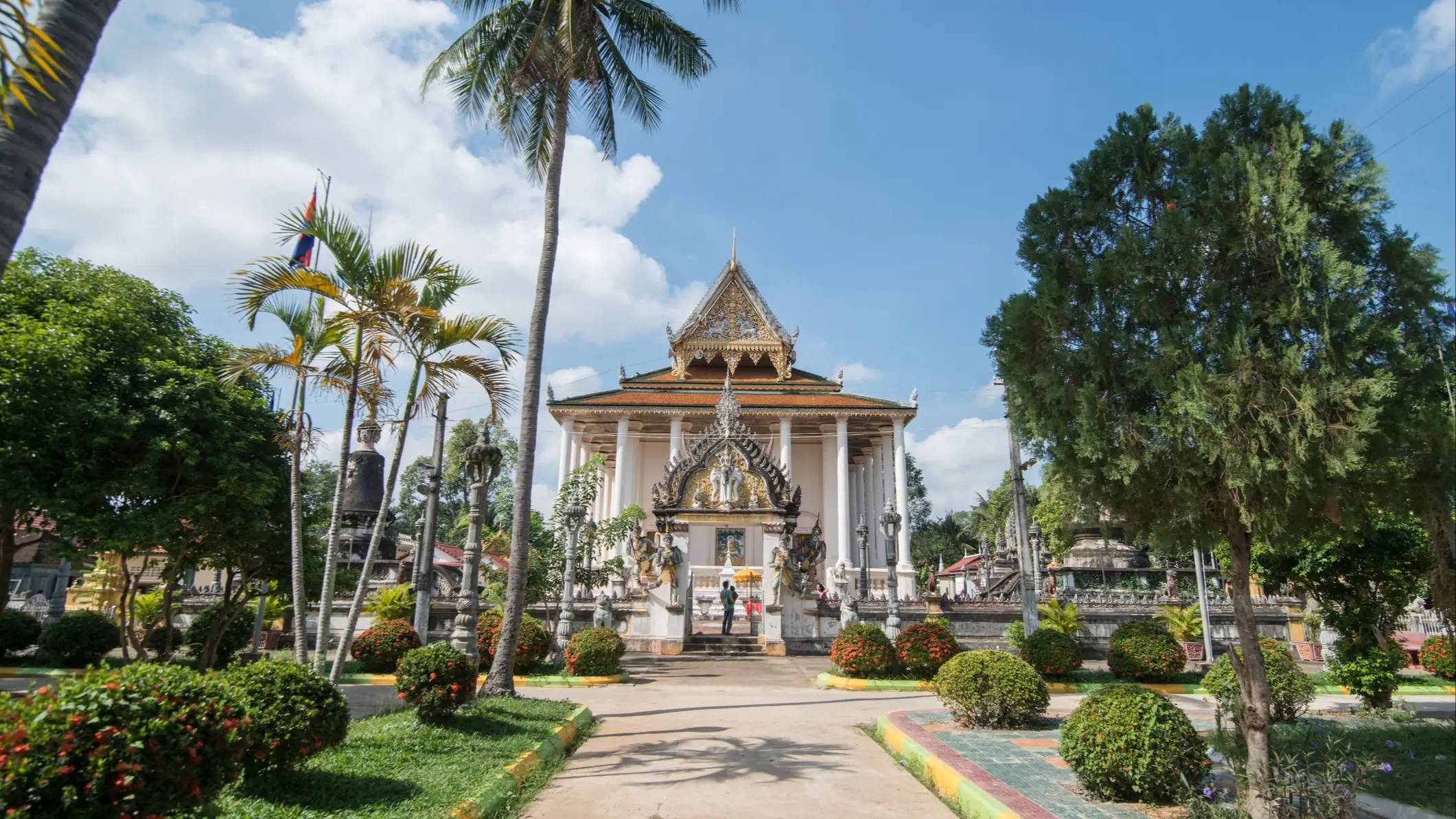 Battambang, temple, Cambodge Un temple traditionnel à Battambang, orné de décorations raffinées et surmonté d'un toit pointu, se dresse au milieu de jardins luxuriants, de palmiers et d'arbustes bien entretenus, sous un ciel d'un bleu éclatant parsemé de quelques nuages. Deux personnes se tiennent à l'entrée du temple.