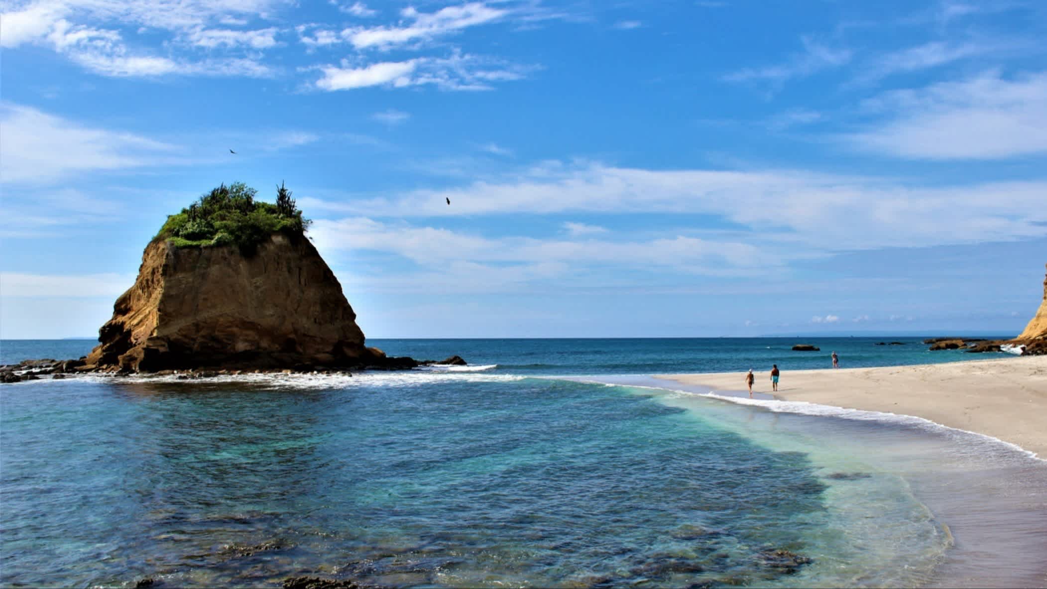 Der Strand Playa Los Frailes, Provinz Manabí, Ecuador bei Sonnenschein und mit Blick auf den hellen Sand sowie einer kleinen vorgelagerten Insel und zwei Menschen die am Strand spazieren.