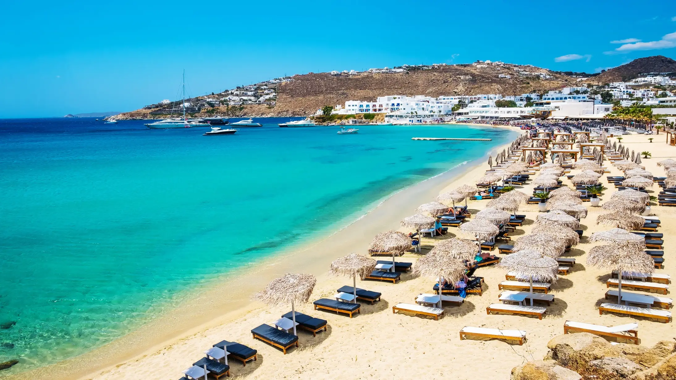 Transats avec des parasols sur le sable blanc au bord de l'eau turquoise, à Mykonos, en Grèce.
