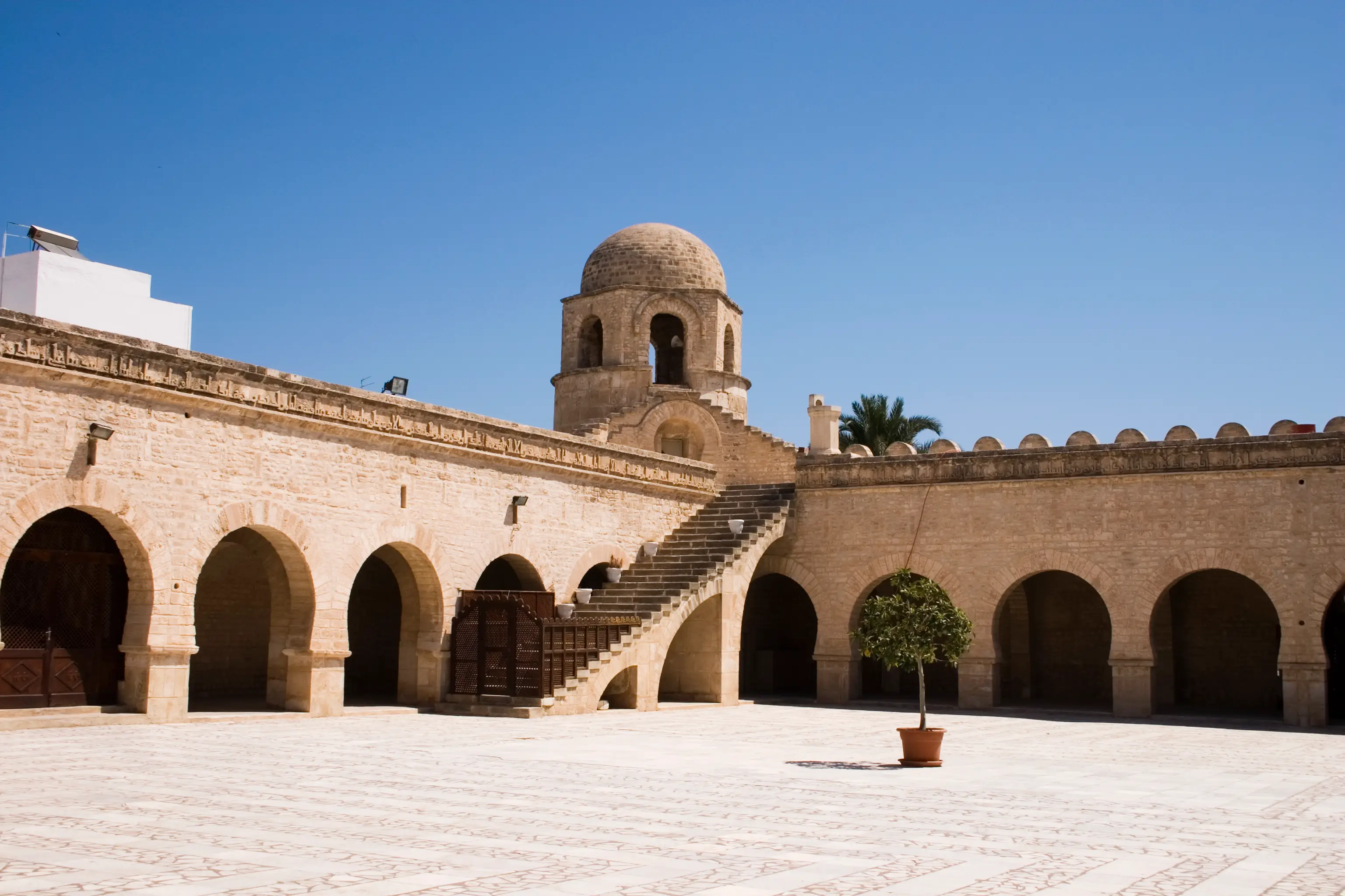 Historical mosque with sandstone walls, arcades, and domed tower under blue sky, small tree in the courtyard.
