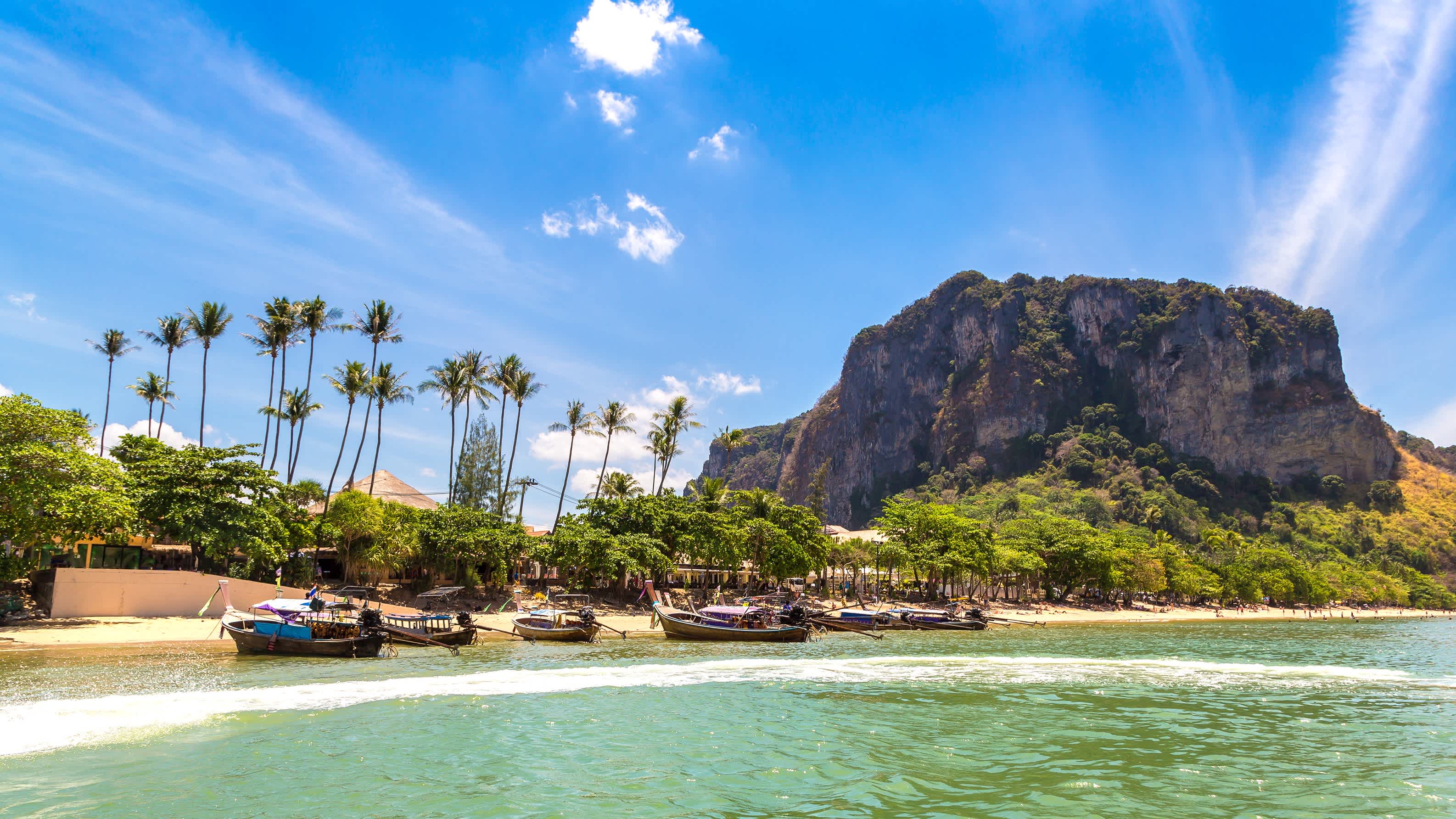 Bateaux sur l'eau claire au bord du sable doré de la plage tropicale d'Ao nang à Krabi, en Thaïlande avec de la végétation et des rochers en arrière plan