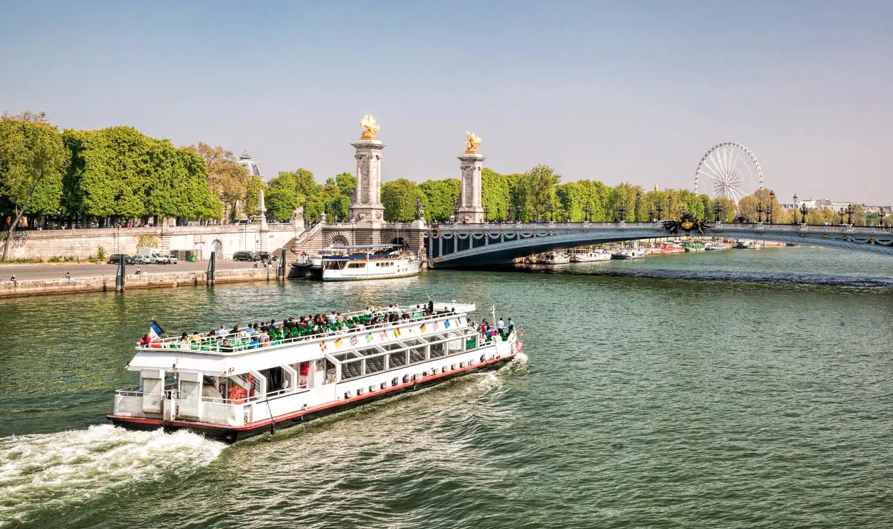 Tourist boat cruising on the Seine River in Paris with Pont Alexandre III bridge and ferris wheel visible in background.