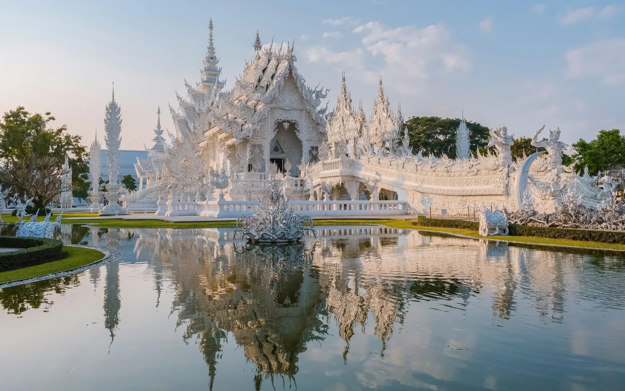 Temple blanc de Wat Rong Khun en Thaïlande se reflétant dans l'eau, architecture ornée sous un ciel bleu clair.