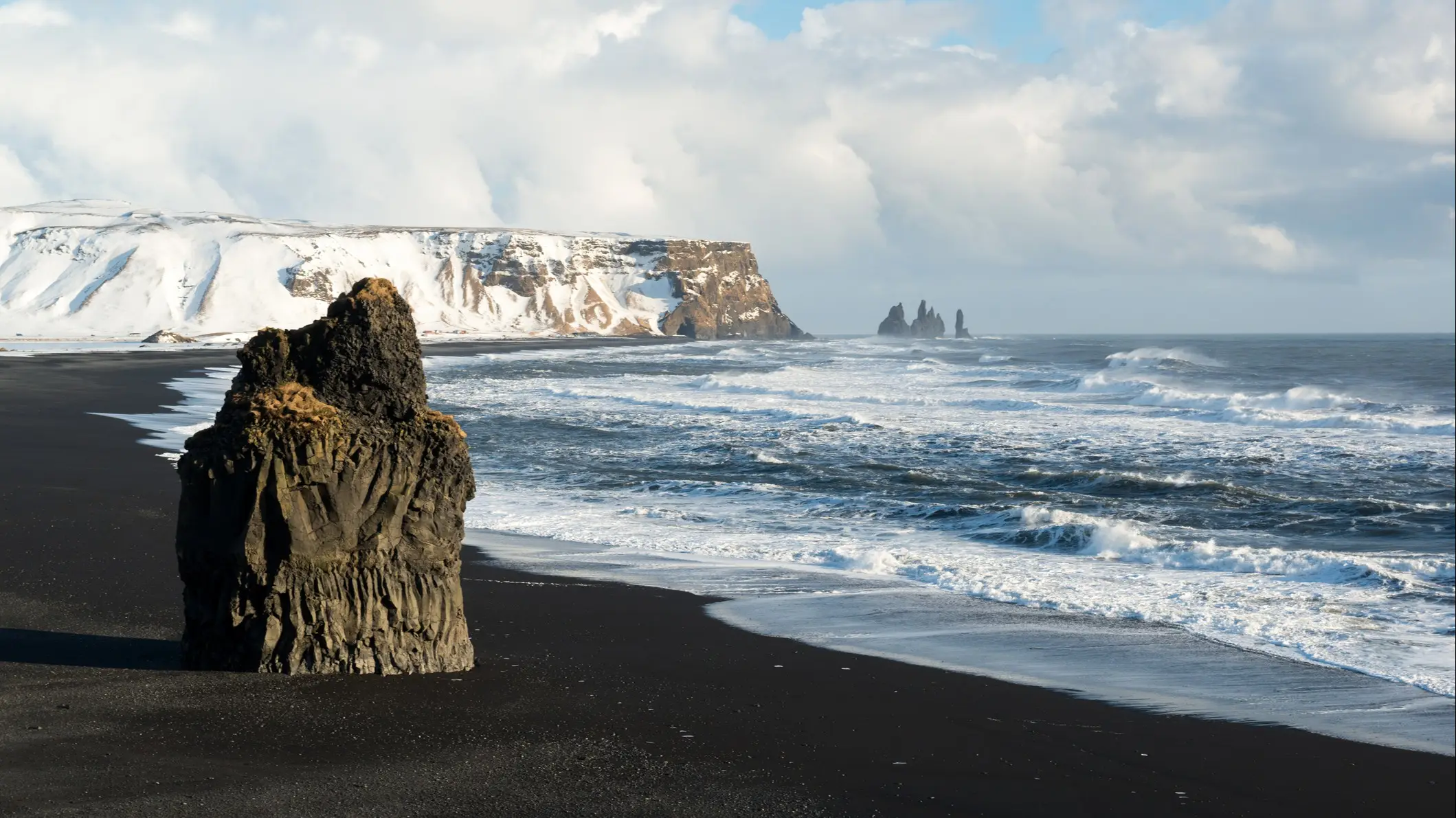Plage de sable noir avec des falaises enneigées en arrière-plan, en Islande.