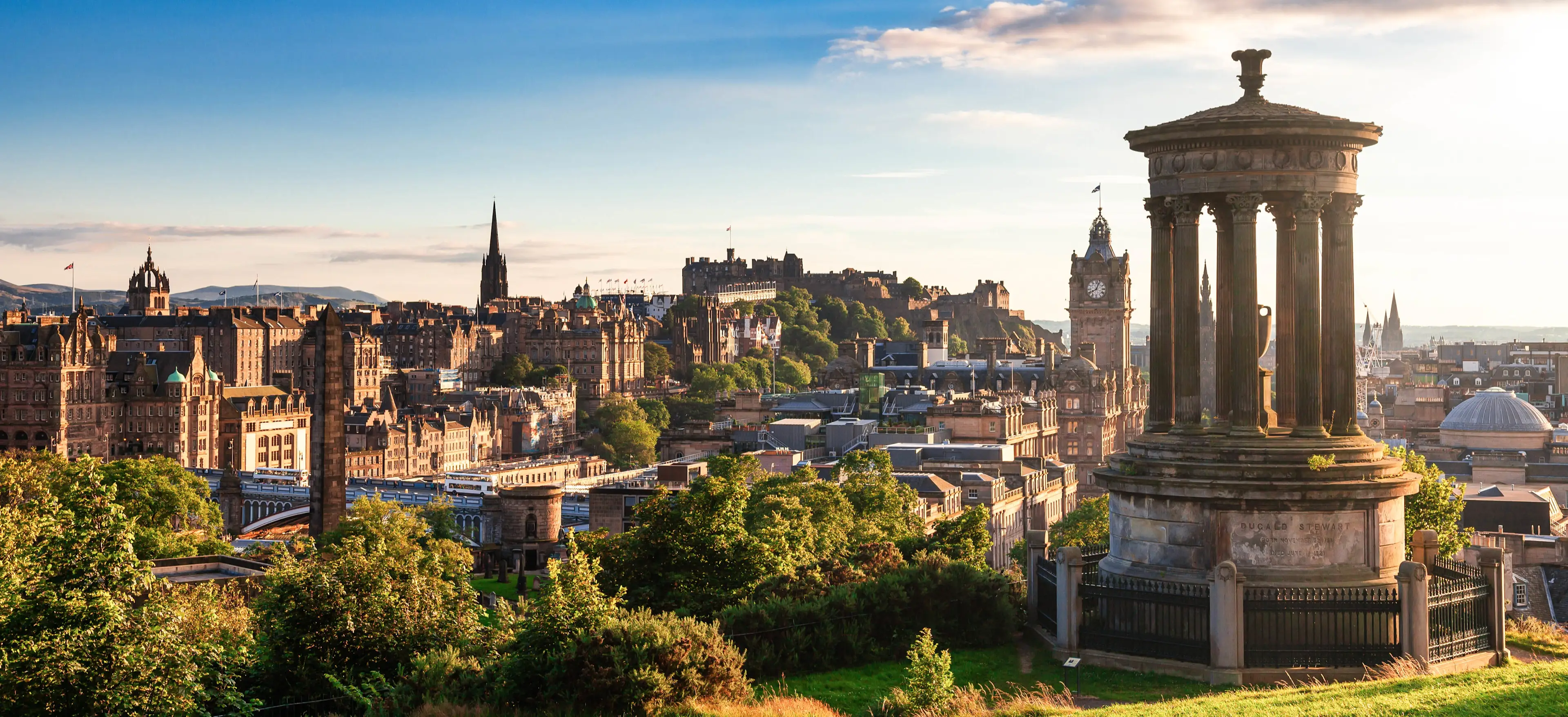 Edinburgh skyline with historic castle, clock tower, and Dugald Stewart Monument in golden evening light.