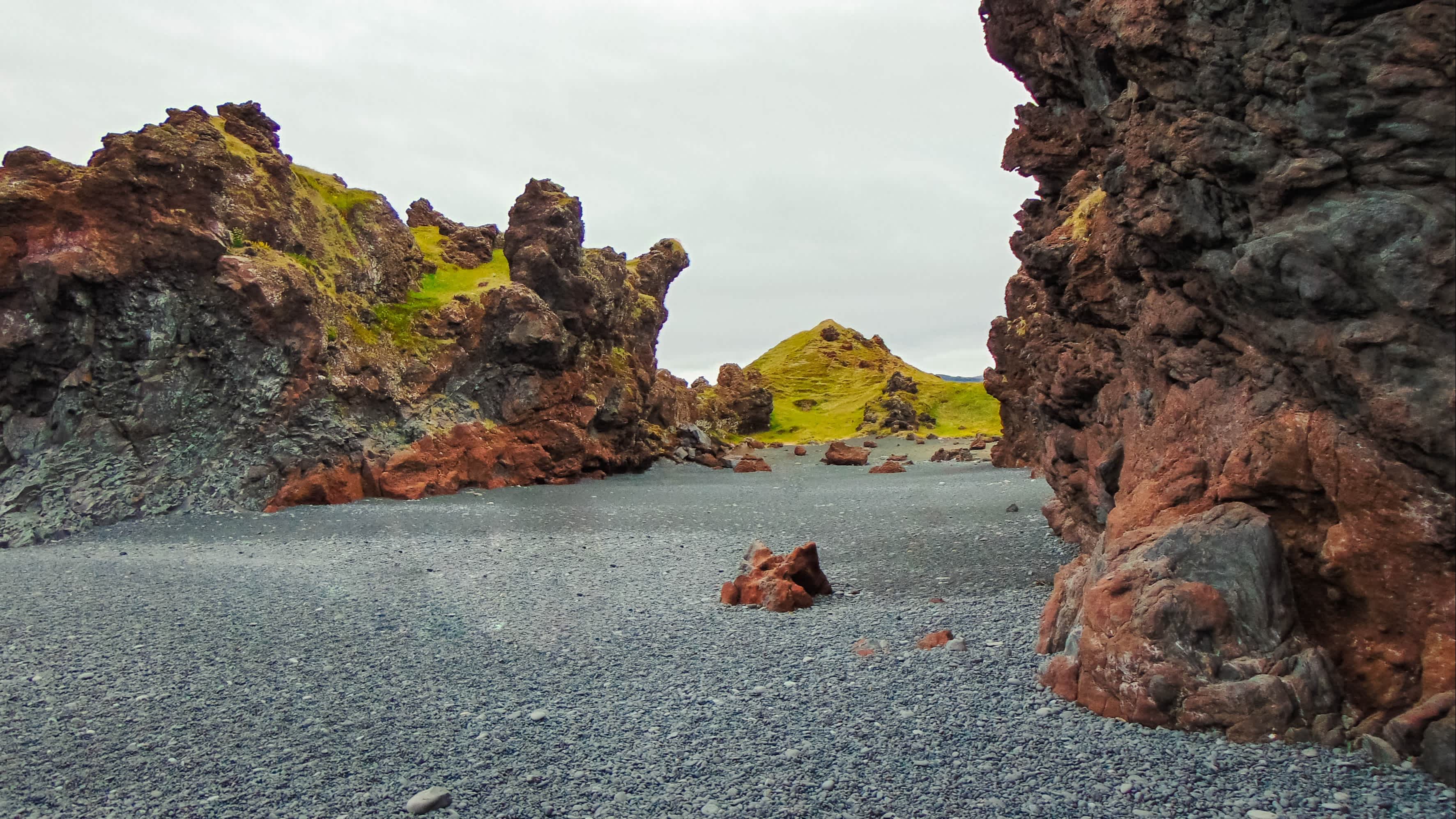 Markanter schwarzer Sandstrand von Djúpalónssandur auf der Halbinsel Snaefellsnes in Island, eingerahmt von schroffen Felsen bei wolkigem Himmel und mit grünen Hügeln im Hintergrund. 