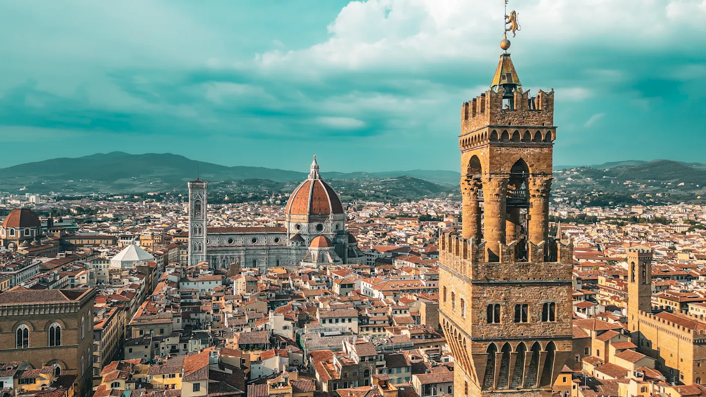 Ein Panoramablick auf Florenz, Italien, mit der roten Kuppel der Kathedrale von Florenz (Duomo), dem hohen Turm des Palazzo Vecchio und historischen Gebäuden mit Terrakotta-Dächern unter einem blauen Himmel mit Wolken.