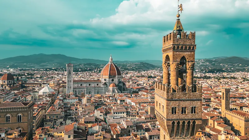 Ein Panoramablick auf Florenz, Italien, mit der roten Kuppel der Kathedrale von Florenz (Duomo), dem hohen Turm des Palazzo Vecchio und historischen Gebäuden mit Terrakotta-Dächern unter einem blauen Himmel mit Wolken.