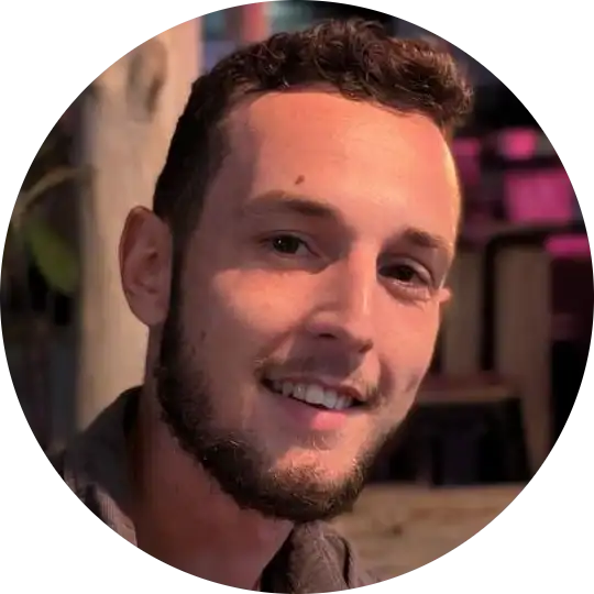 Un jeune homme aux cheveux courts bouclés et à la barbe taillée sourit à la caméra, assis dans une pièce faiblement éclairée, entouré de plantes et avec un fond flou.