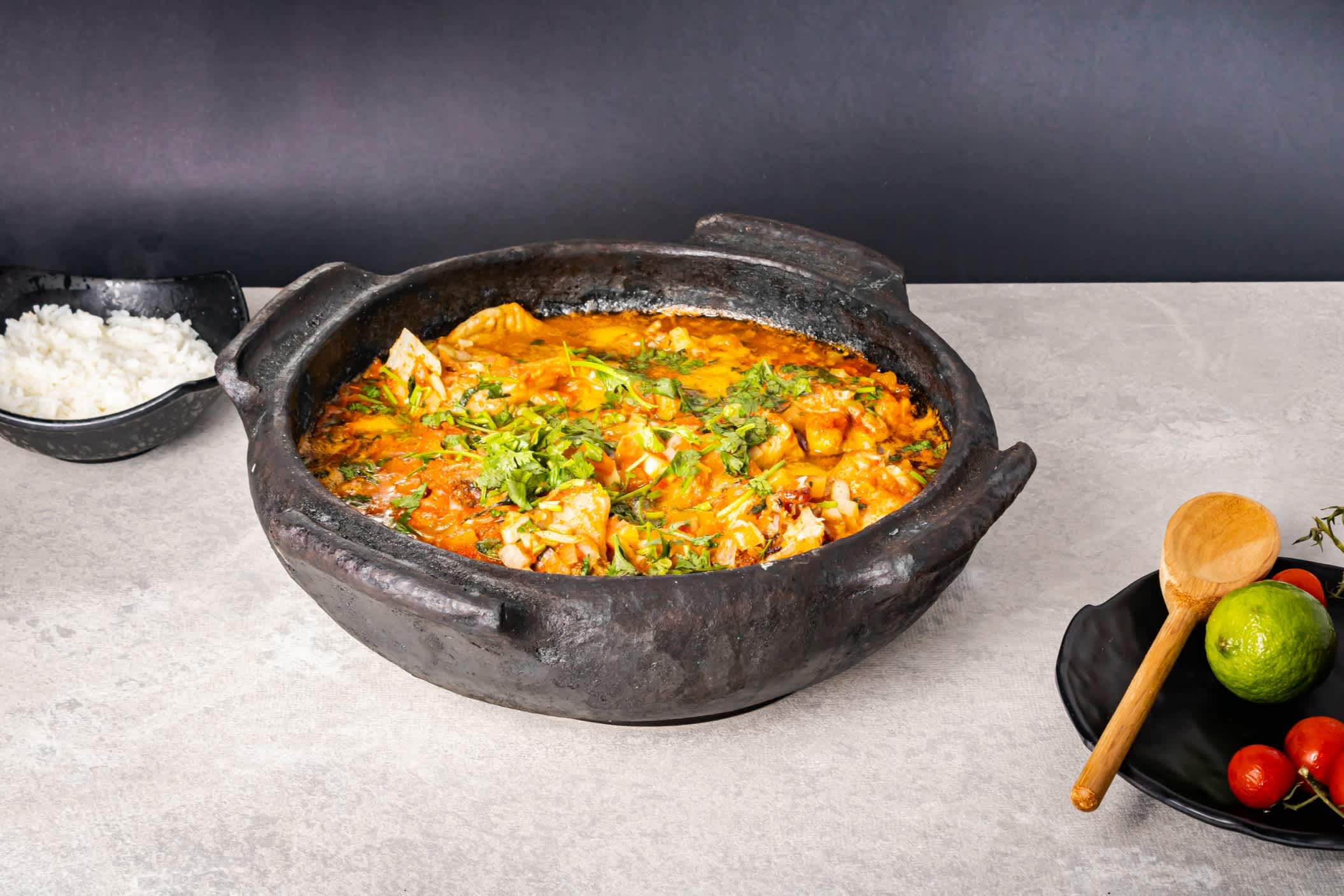 Délicieuse moqueca de poisson brésilienne avec tomates, oignons, huile d'olive, coriandre et graines d'orocum. Le plat est présenté dans un pot en terre cuite sur une table blanche et un fond noir.