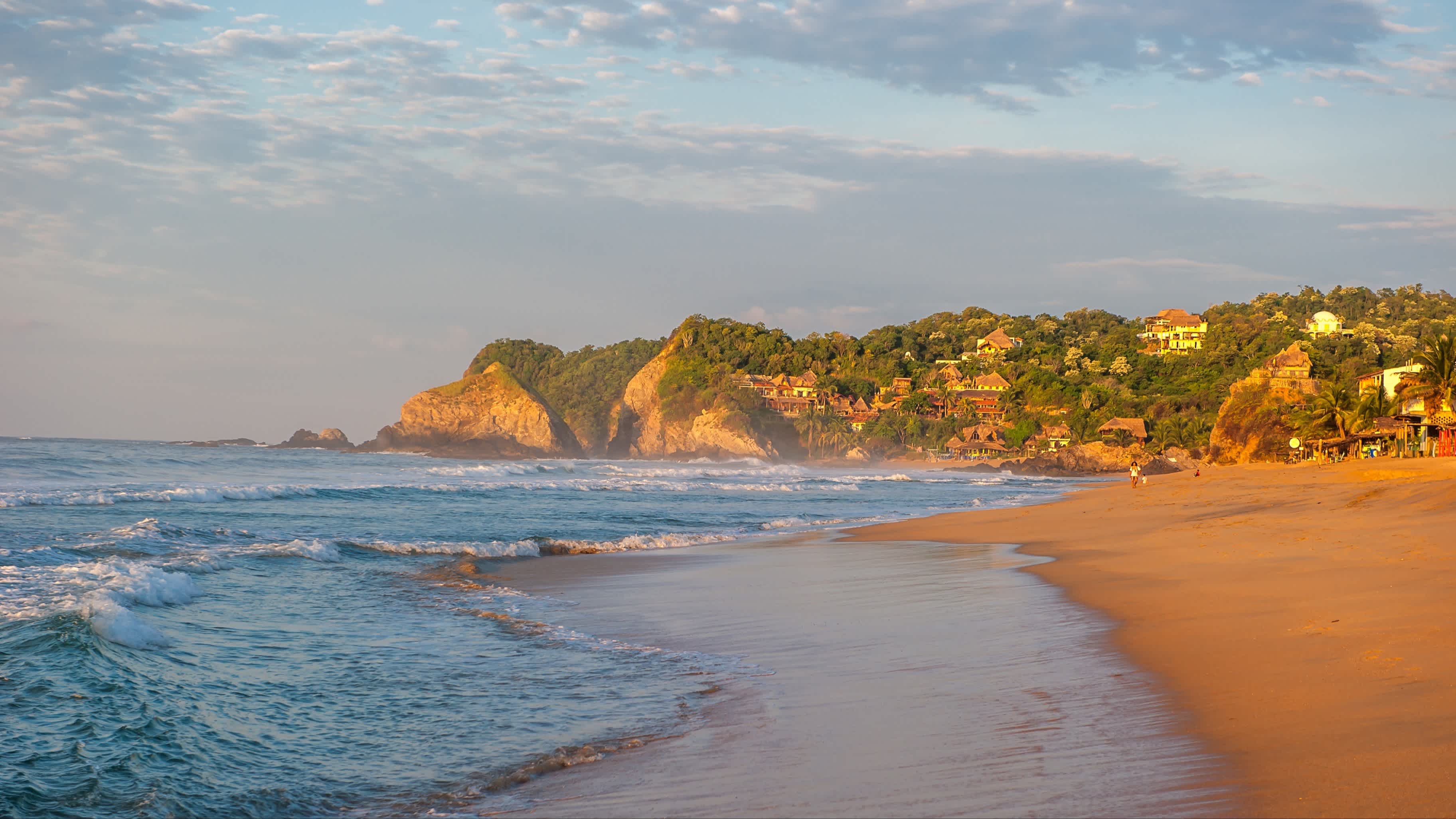 Zipolite Strand bei Sonnenaufgang, Mexiko und mit Blick auf das Meer sowie Häuser an der Küstenlinie und Felsen.