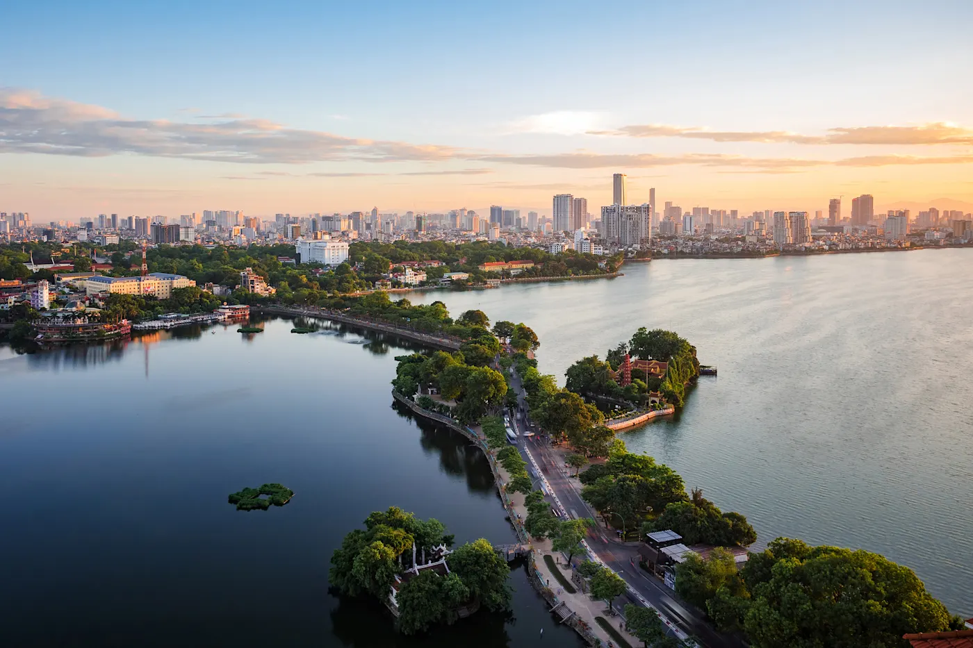 Hanoi Luftaufnahme einer Stadt mit einem großen See, einer von Bäumen gesäumten Halbinsel und Straßen bei Sonnenuntergang. Im Hintergrund sind hohe moderne Gebäude bei teilweise bewölktem Himmel zu sehen.