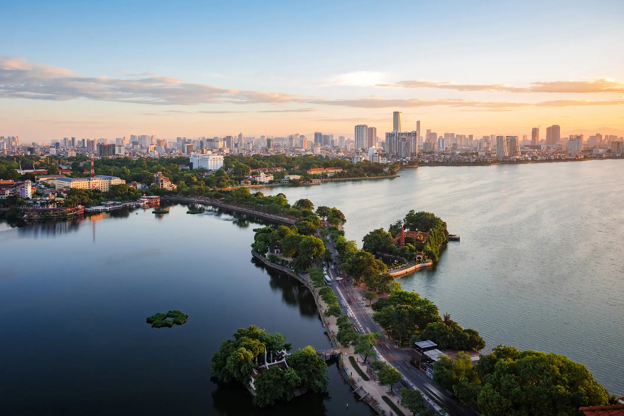 Hanoi Luftaufnahme einer Stadt mit einem großen See, einer von Bäumen gesäumten Halbinsel und Straßen bei Sonnenuntergang. Im Hintergrund sind hohe moderne Gebäude bei teilweise bewölktem Himmel zu sehen.