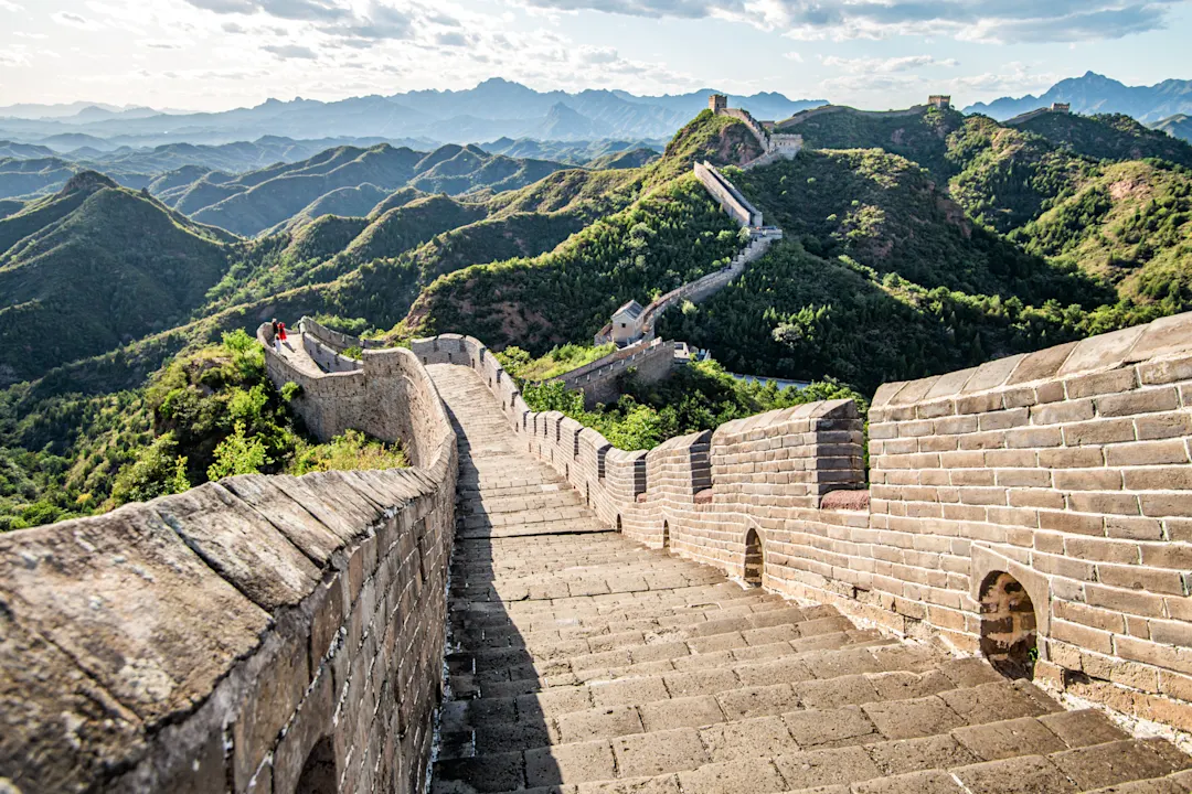 Die Chinesische Mauer schlängelt sich durch grüne Berglandschaft unter blauem Himmel mit weißen Wolken.