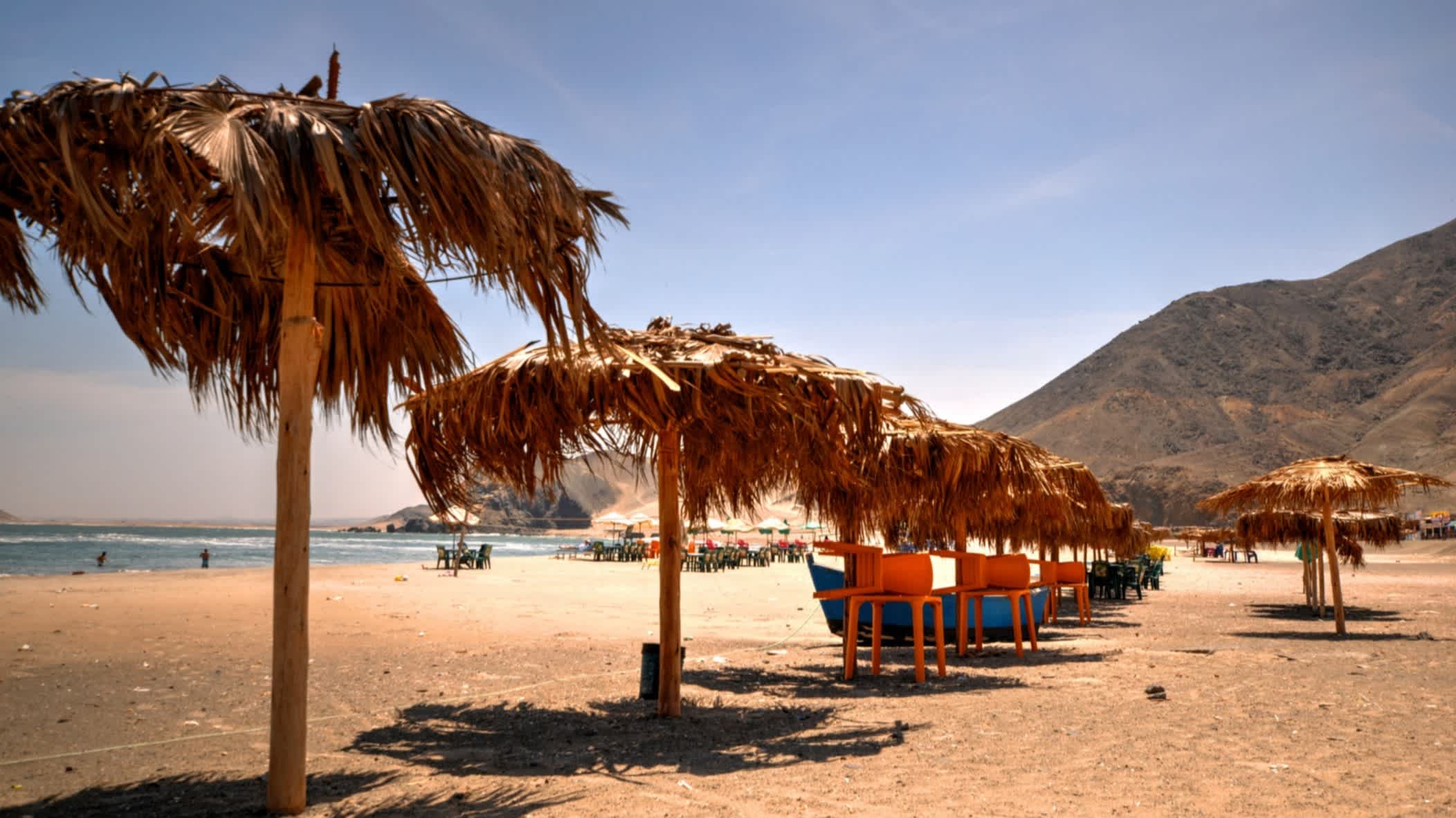 Der Strand Playa Vesique in Ancash, Peru mit traditionellen Sonnenschirmen aus Stroh im Bild und Blick auf den goldenen Sand, die Berge im Hintergrund und das Meer mit Badenden darin.
