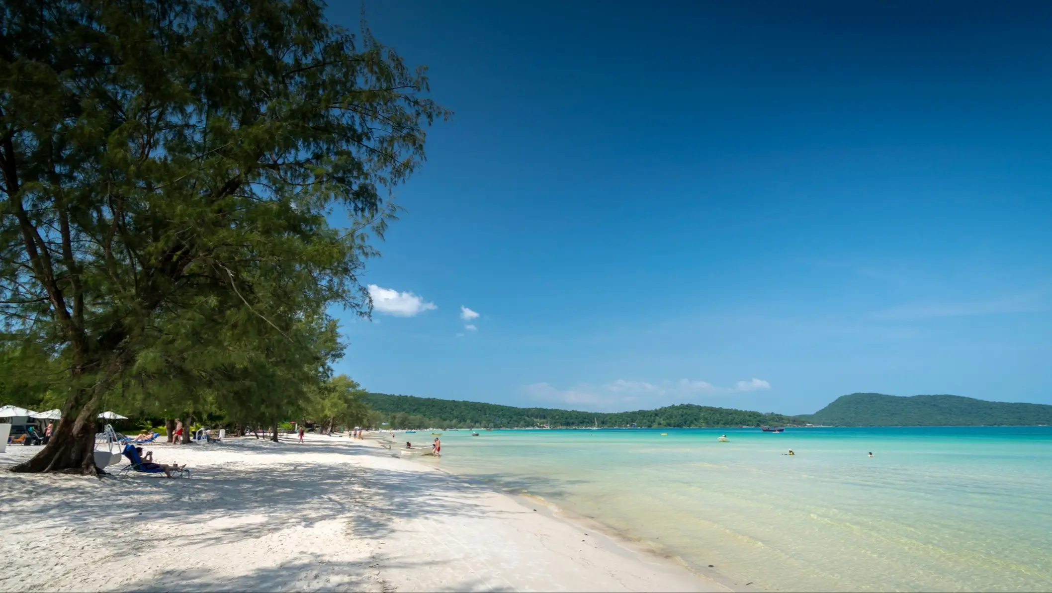 Tropischer Strand mit weißem Sand, türkisblauem Wasser, grünen Bäumen und bewaldeten Hügeln unter klarem Himmel.