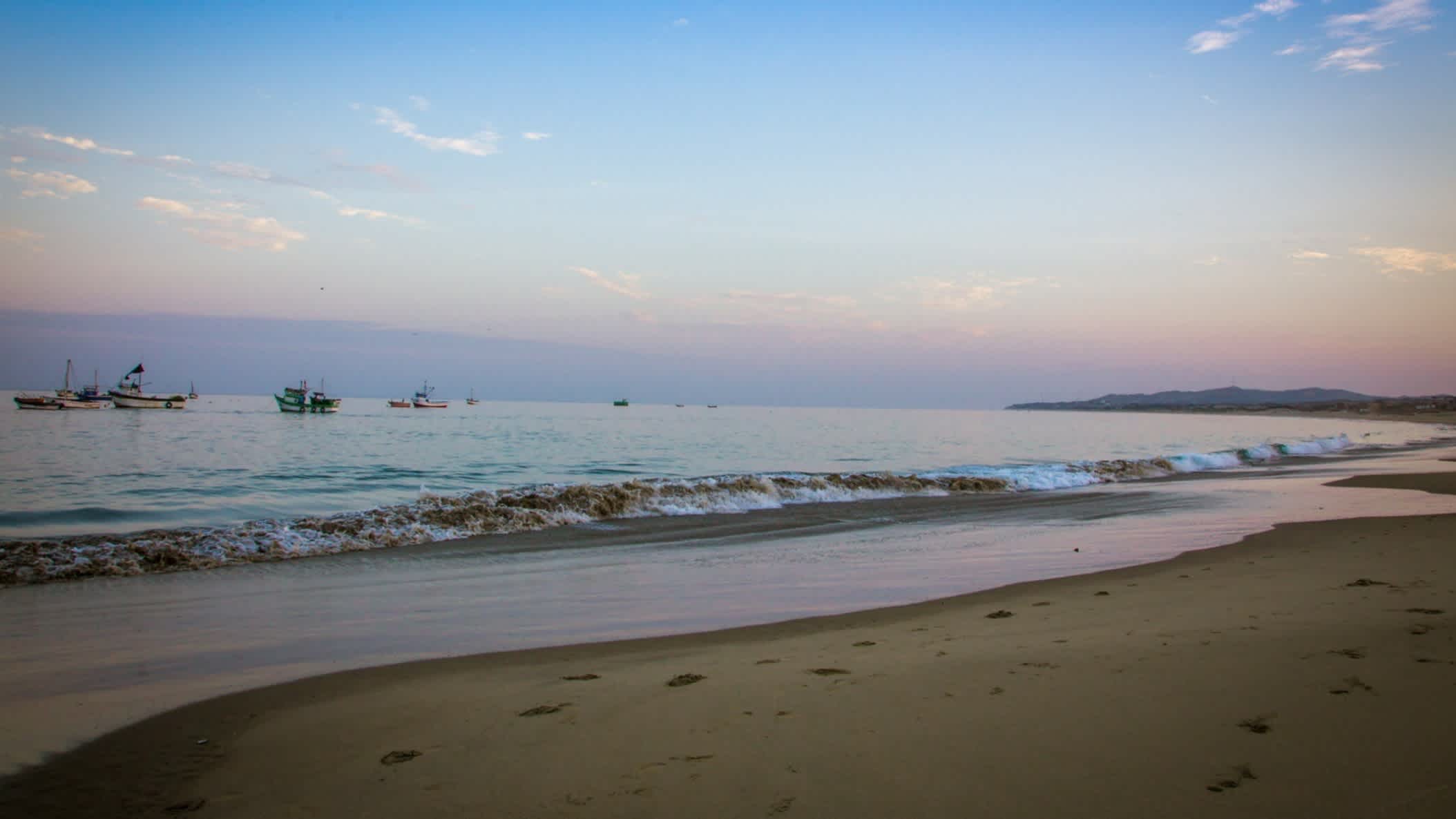 Der Strand Playa Los Organos in Mancora, Peru bei Sonnendämmerung sowie mit Booten im seichten Wasser und dem goldenen Sand im Vordergrund.