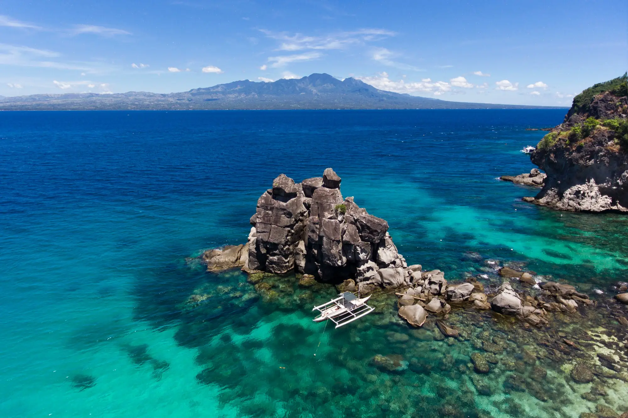 Türkisblaues Meer mit Felsformation und kleinem Boot, im Hintergrund Bergkette unter blauem Himmel mit weißen Wolken.
