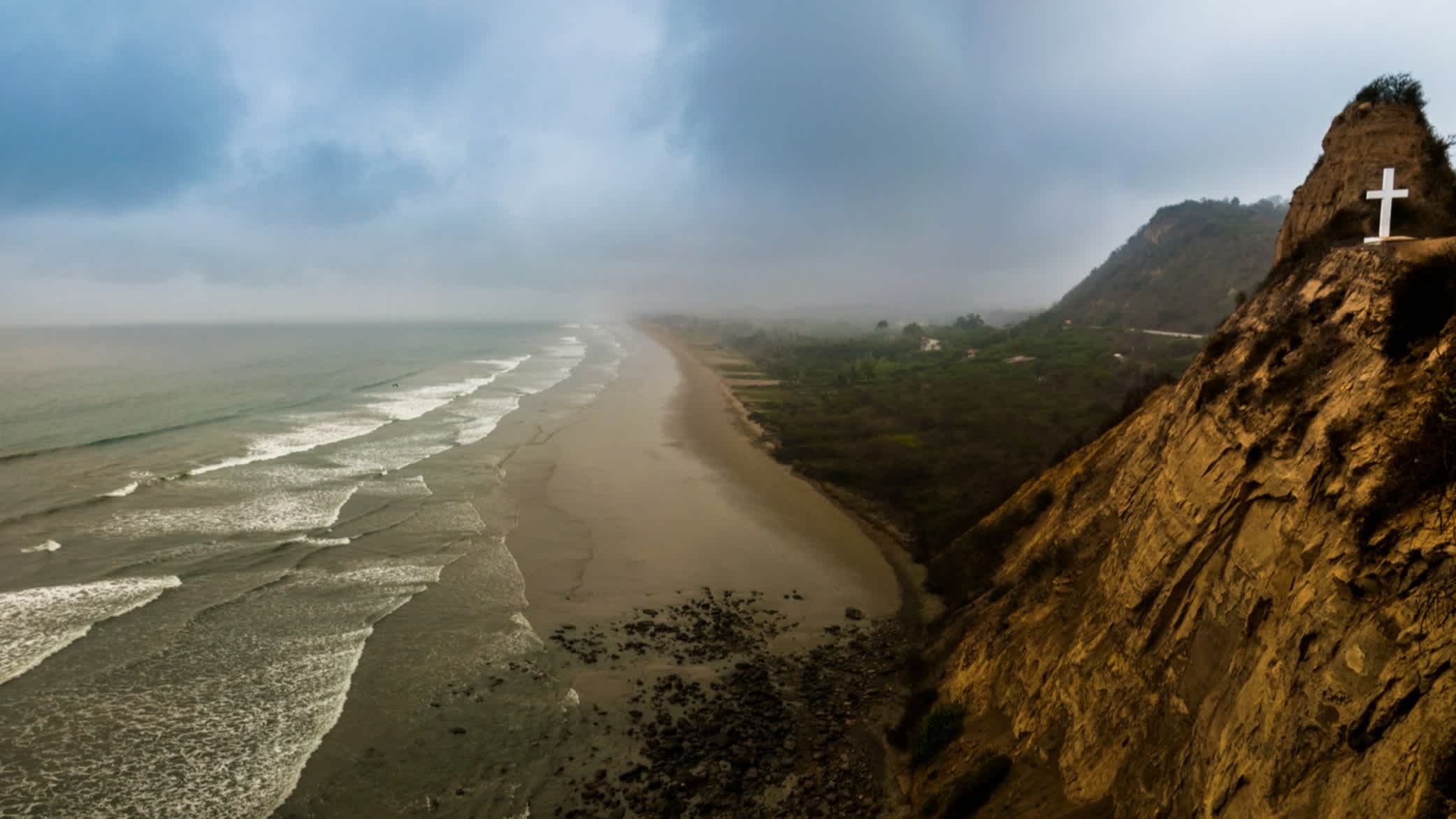 Der Strand von Montañita, Provinz Santa Elena, Ecuador bei bewölktem Himmel und mit Ausblick auf das weite Meer mit Wellen sowie dem Gebirge.