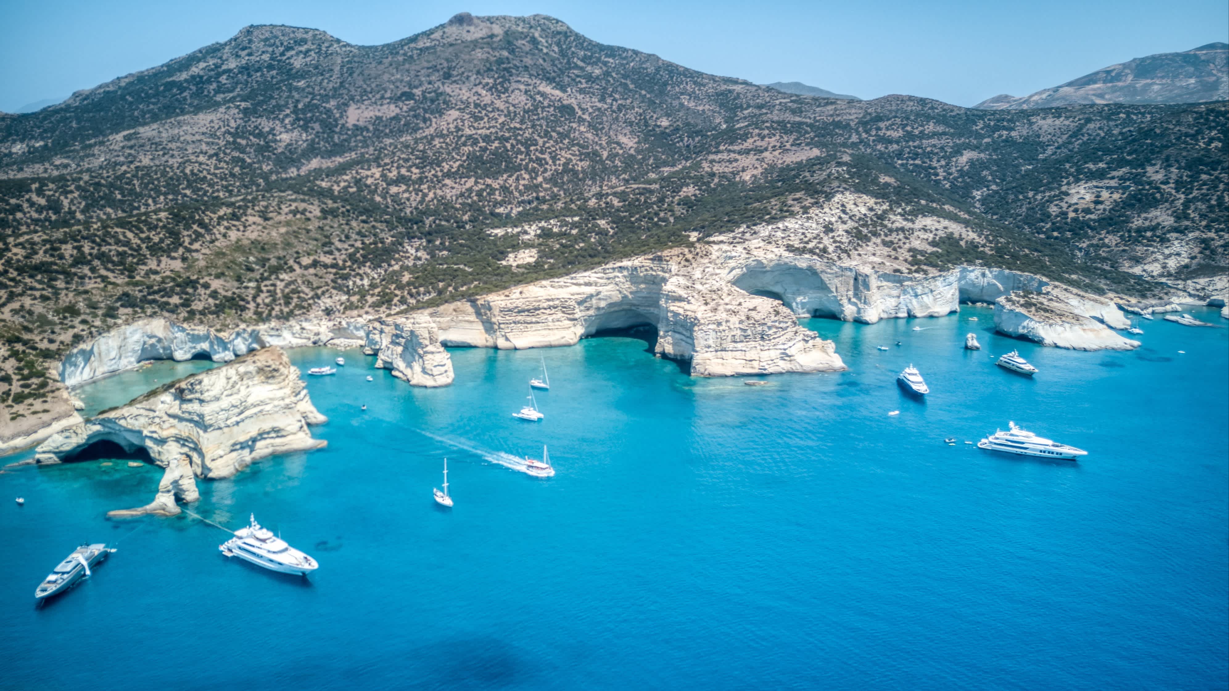 Eine Luftaufnahme der Kleftiko-Bucht auf der Insel Milos, Griechenland mit Blick auf die Küstenlinie und das blaue Meer mit Booten im Wasser sowie Bergen im Hintergrund.