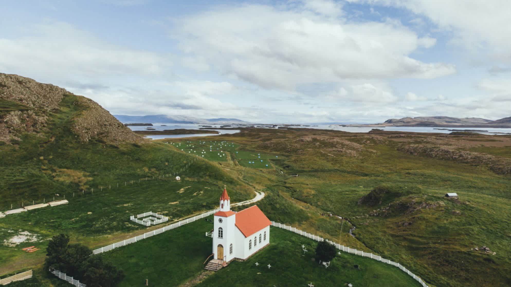 Malerische Luftaufnahme einer Kirche in Island, umgeben von grünen Wiesen, Bergen, Klippen und Meer am Horizont, bei wolkigem Wetter. 