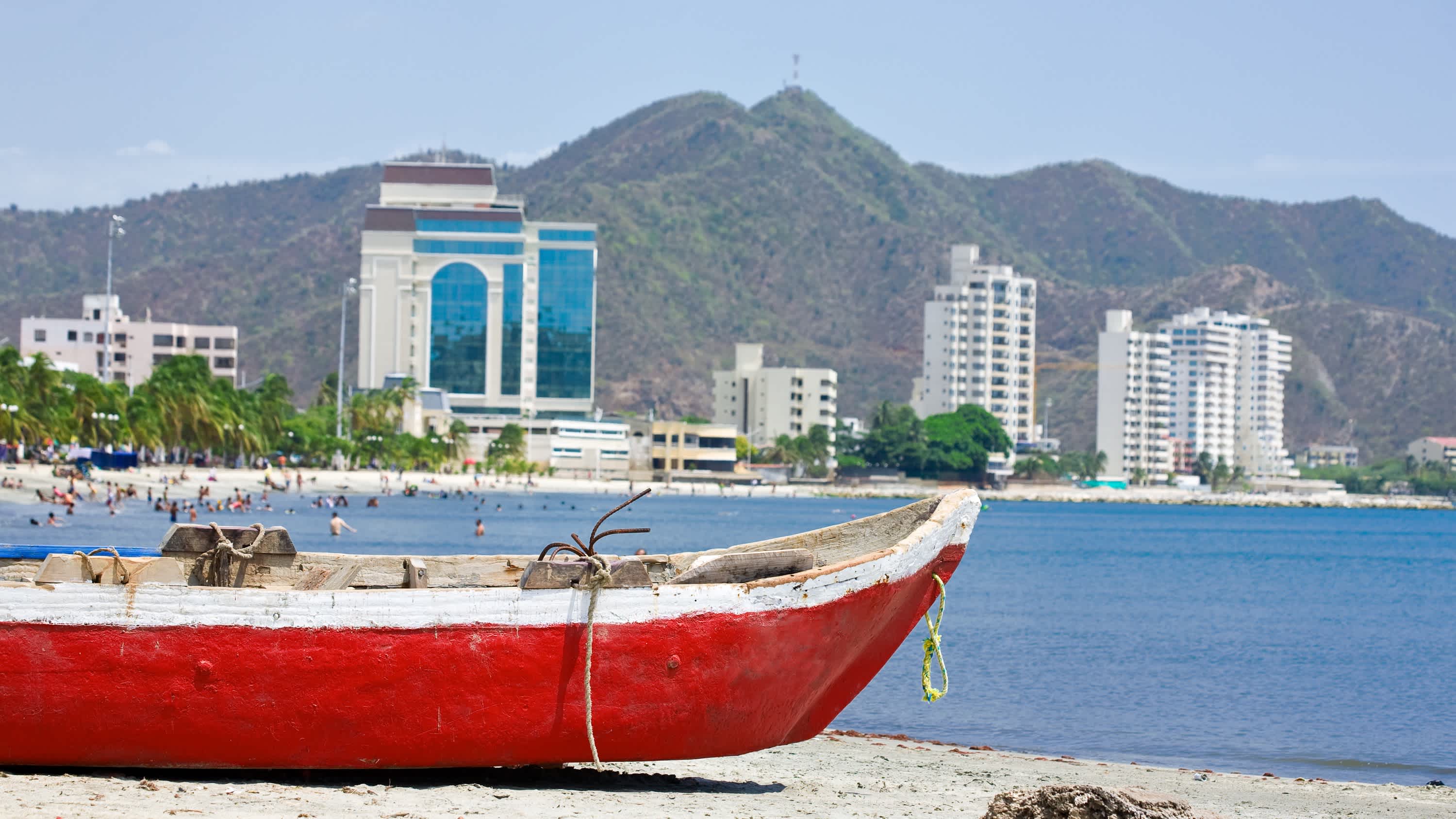  Playa Rodadero in Santa Marta, Kolumbien mit einem roten Boot im Vordergrund sowie dem Strand, der Bucht und Hochhäusern im Hintergrund.