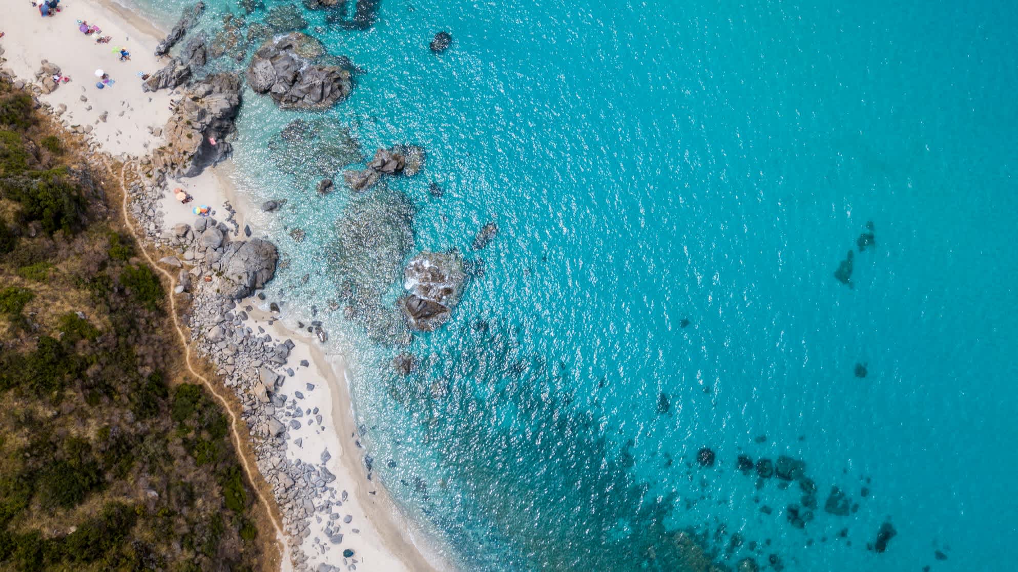 Luftaufnahme des Strandes in Zambrone, Kalabrien, mit türkisblauem Meer, üppiger Vegetation und Felsen.
