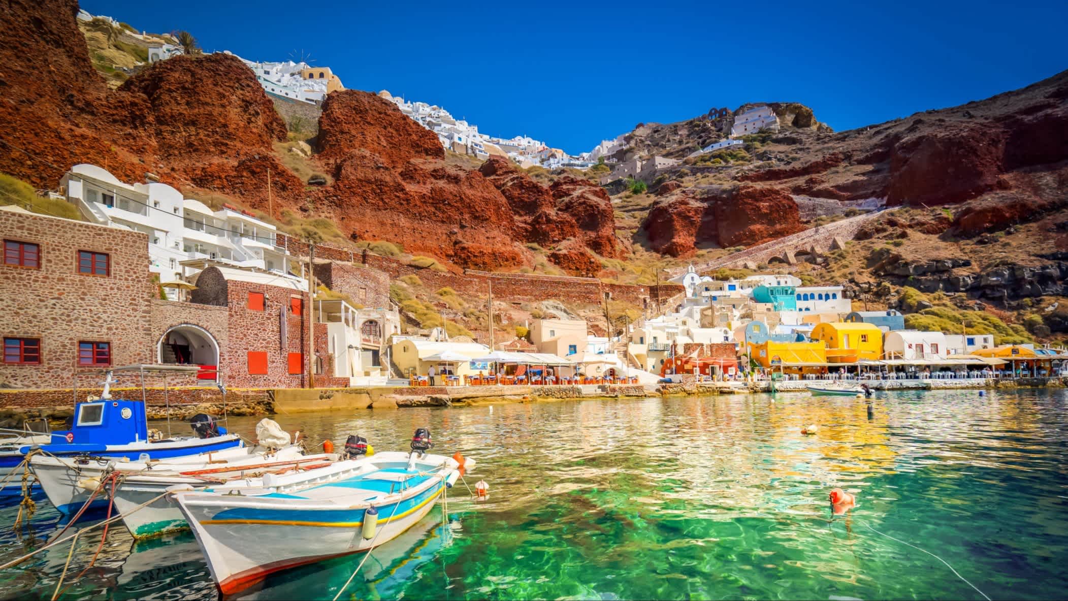Blick zur farbenfrohen Amoudi-Bucht, Santorin, Griechenland mit kristallklarem Wasser sowie Booten im Wasser und Häusern am Rand sowie roten Felsen.