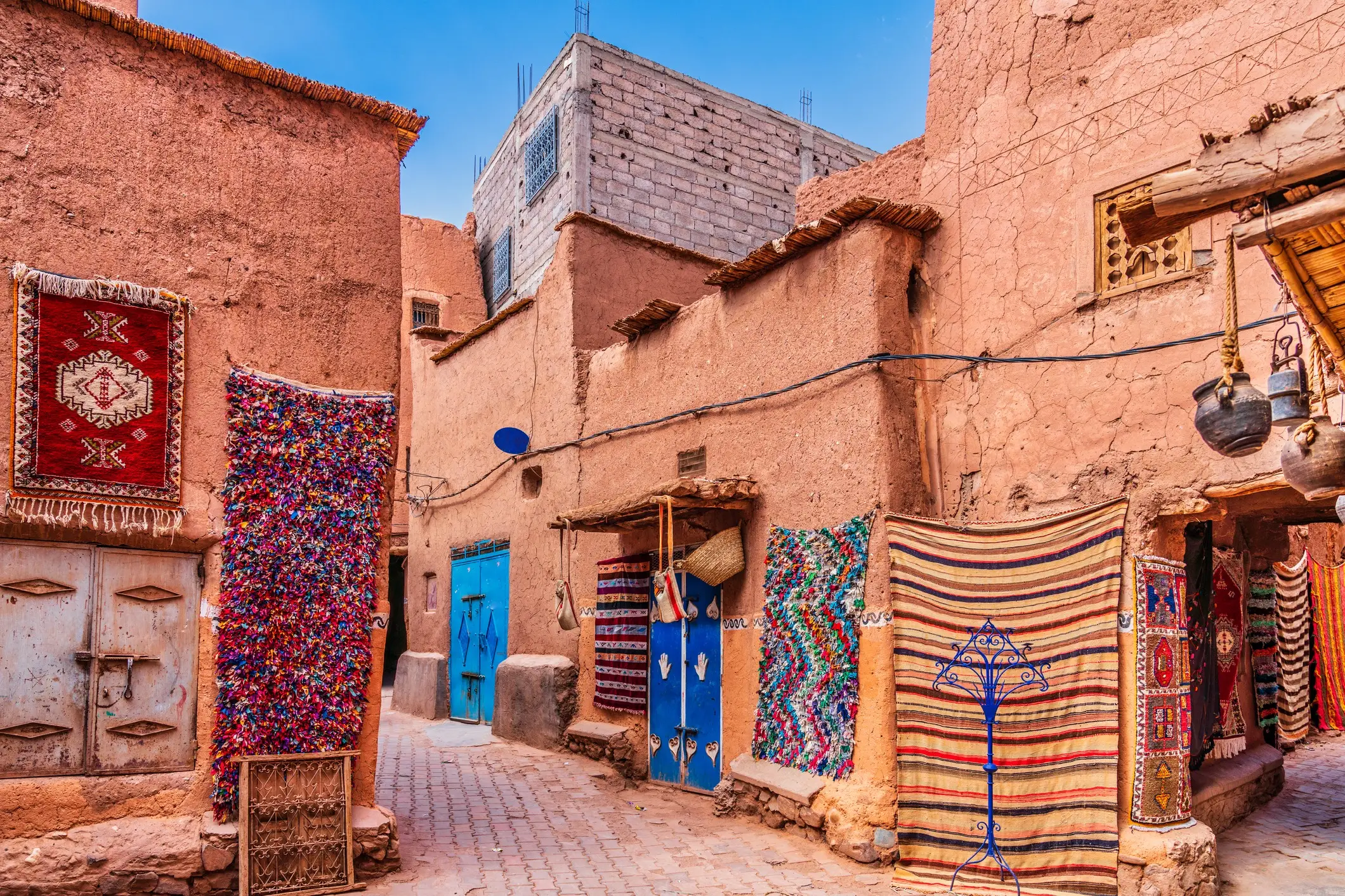 Ruelle colorée dans une médina à Marrakech avec des tapis traditionnels et des portes bleues sur des murs en terre.