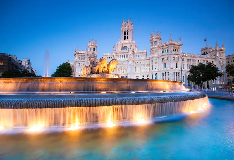 Madrid Der beleuchtete Cibeles-Brunnen mit kaskadenförmigem Wasser vor dem großen Palacio de Cibeles in der Abenddämmerung in Madrid, Spanien, mit einem klaren blauen Himmel im Hintergrund.