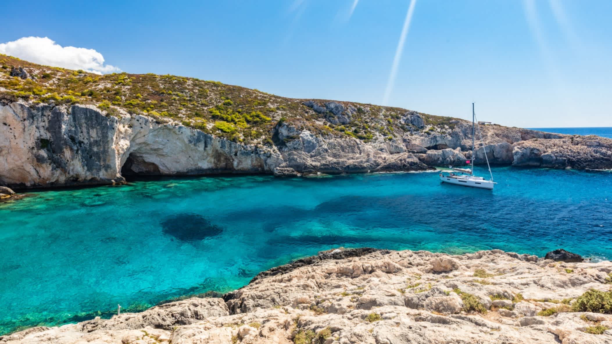 Kristallklares Wasser mit Höhlen am Strand von Limnionas, Zakynthos, Griechenland bei strahlendem Sonnenschein und mit einem Segelboot im Wasser.

